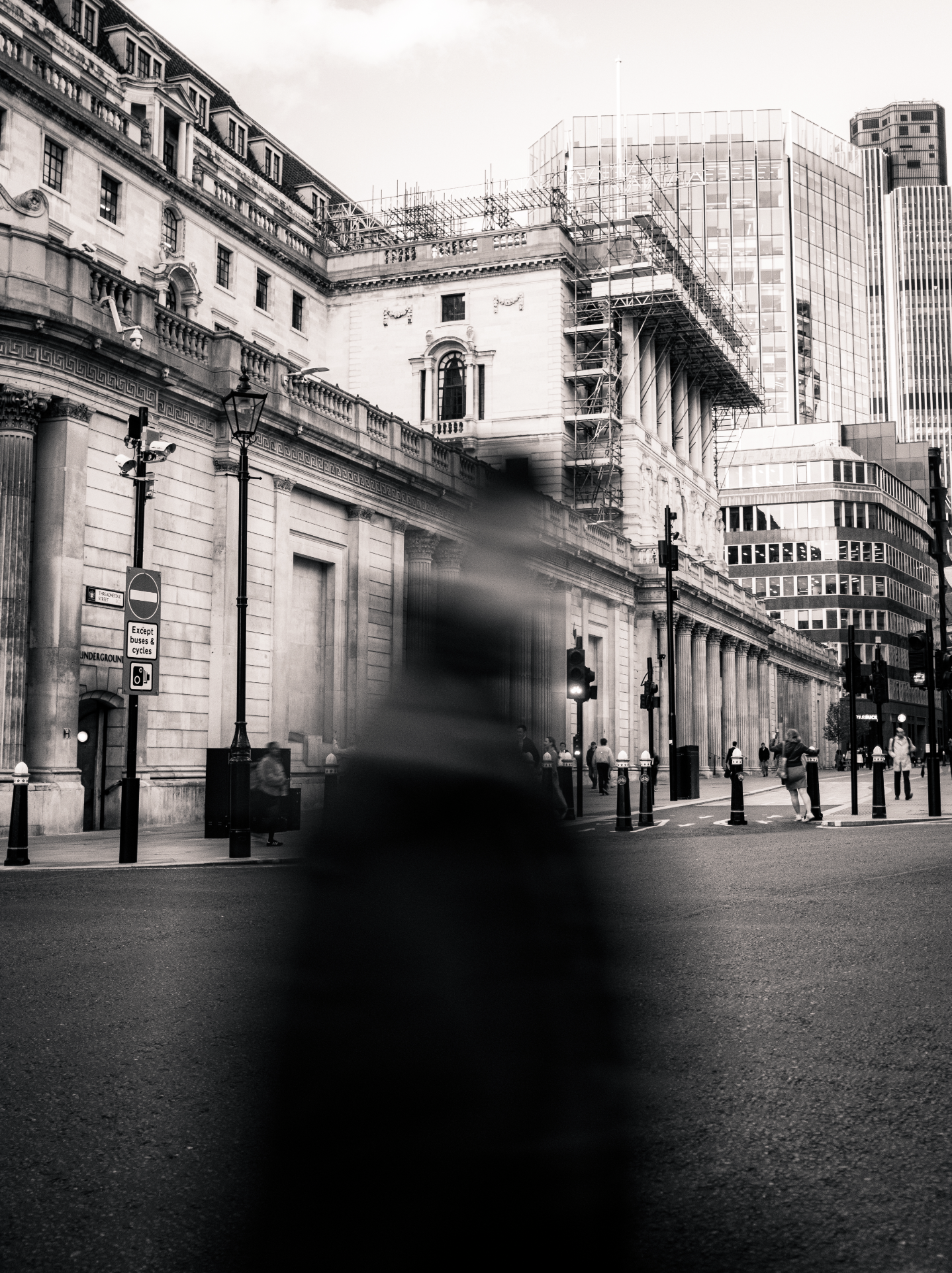 Black and white photo of a city street with tall buildings. Pedestrians walk on the sidewalk, some crossing at the traffic light. A large, ornate building with scaffolding on its upper part is in the background. A blurred object is in the foreground.