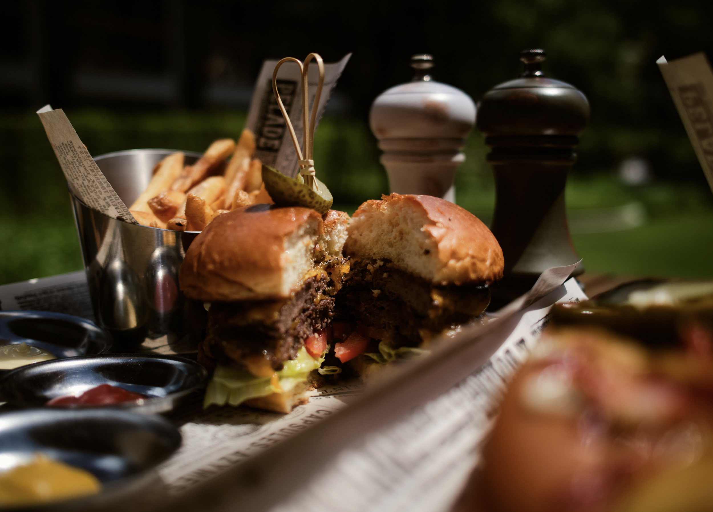 Close-up of a burger with a bun, lettuce, tomato, cheese, and a beef patty, with French fries and condiments in the background, outdoors.