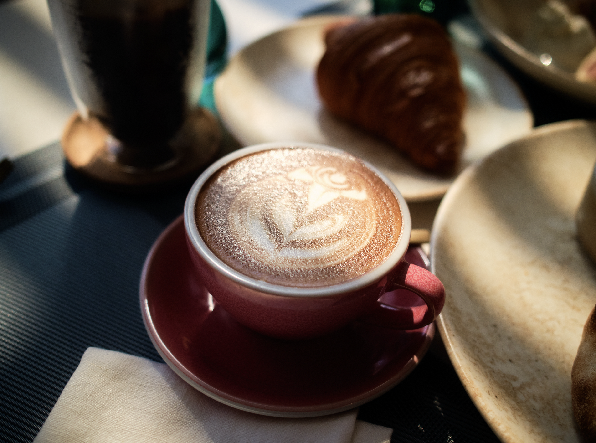 A white cup with a hot beverage topped with foam art, placed on a red saucer, surrounded by pastries on a tray, with a woven placemat underneath.