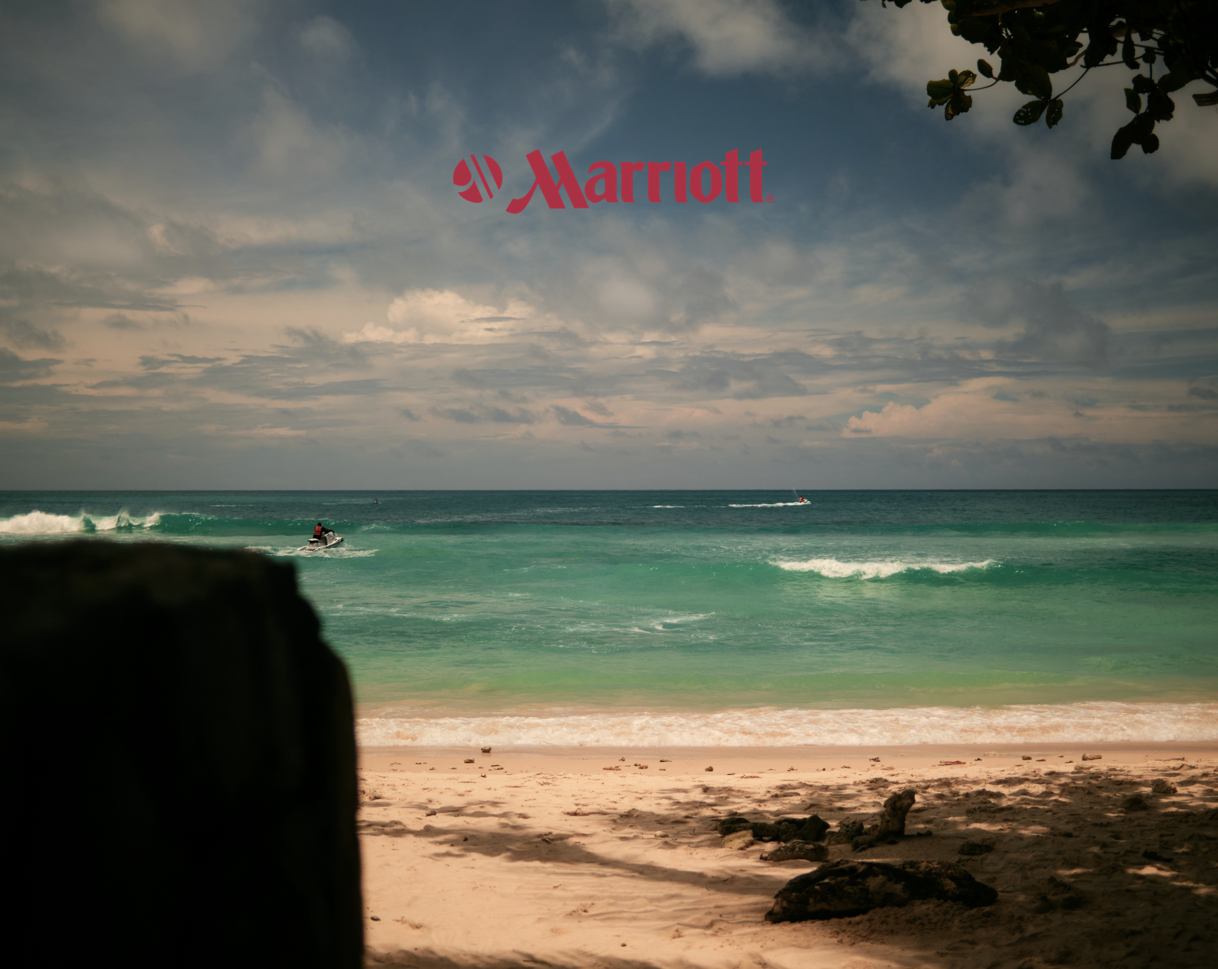 View of a beach seen through a dark opening, with the ocean and cloudy sky in the background, and jet skis riding on the water.