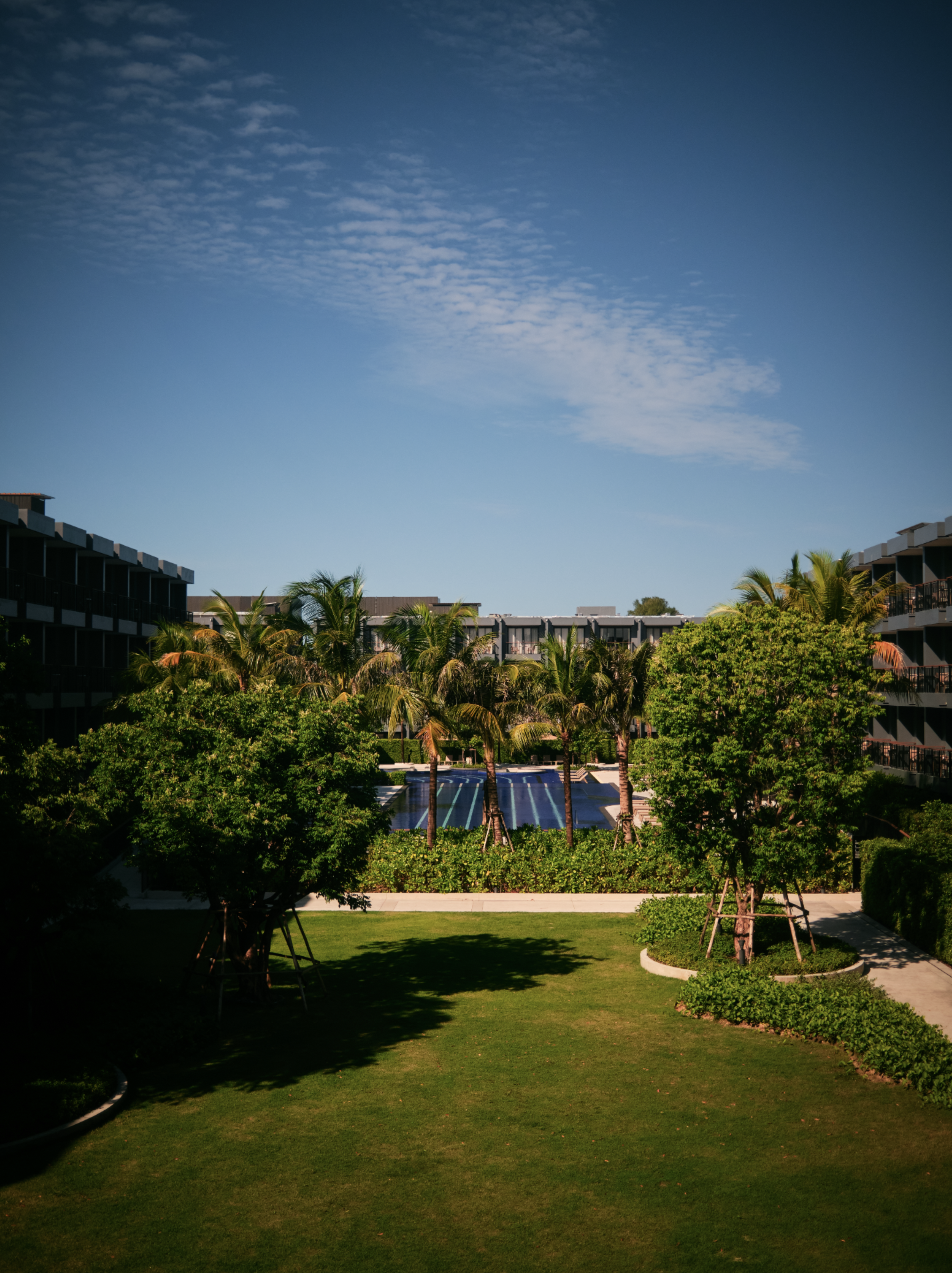 A landscaped garden with lush grass, trees, and shrubs in the foreground. In the background, there is a pool area with lounge chairs, surrounded by mid-rise apartment or hotel buildings. The sky is clear with a few scattered clouds.