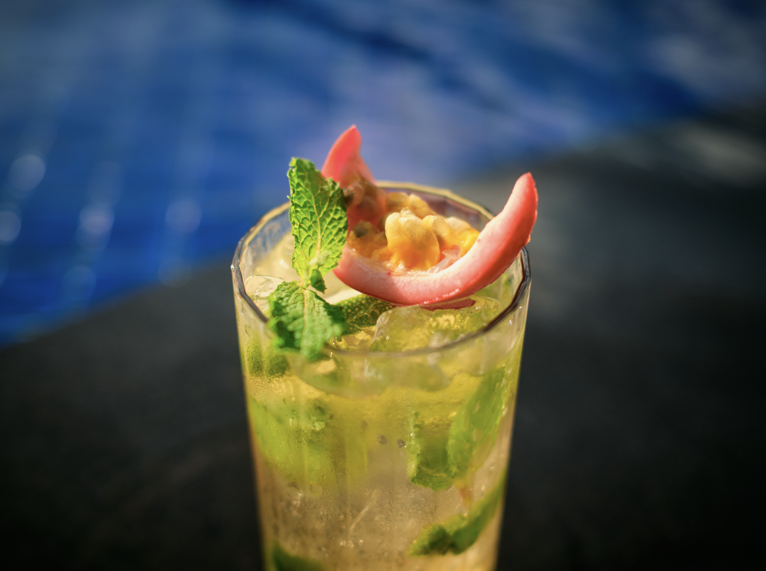 A close-up of a cocktail glass filled with a yellow-green drink, garnished with a slice of passion fruit, mint leaves, and a pink orchid flower, set against a blurred blue background.