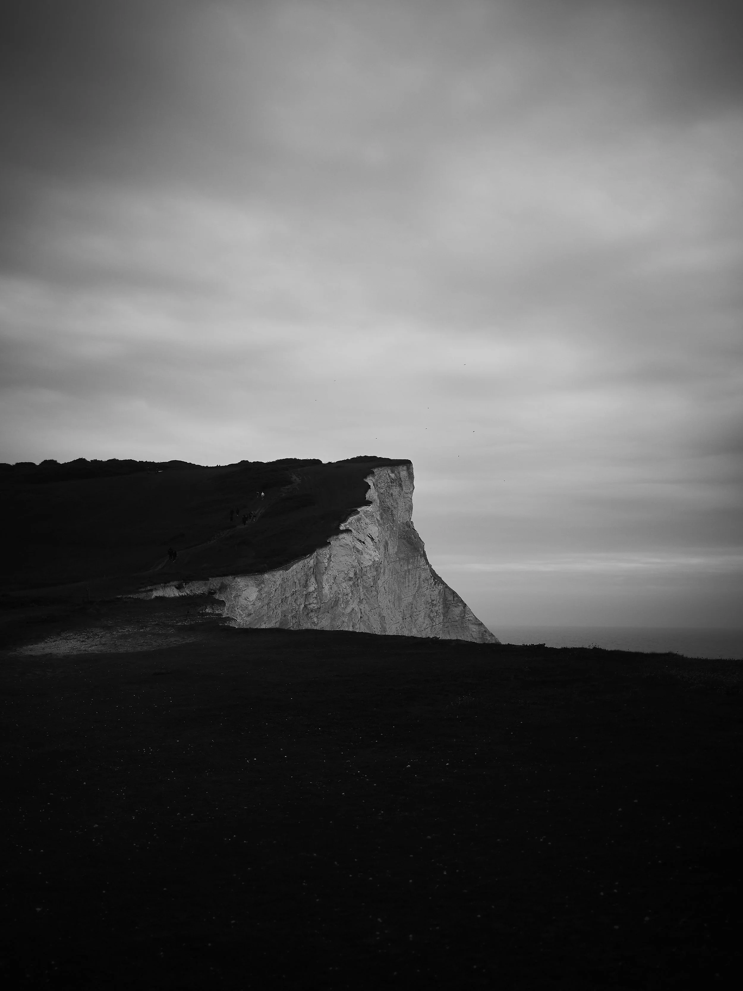 Black and white photograph of a rugged coastal cliff with some small figures walking along the top, under a cloudy sky, possibly near sunset or sunrise.