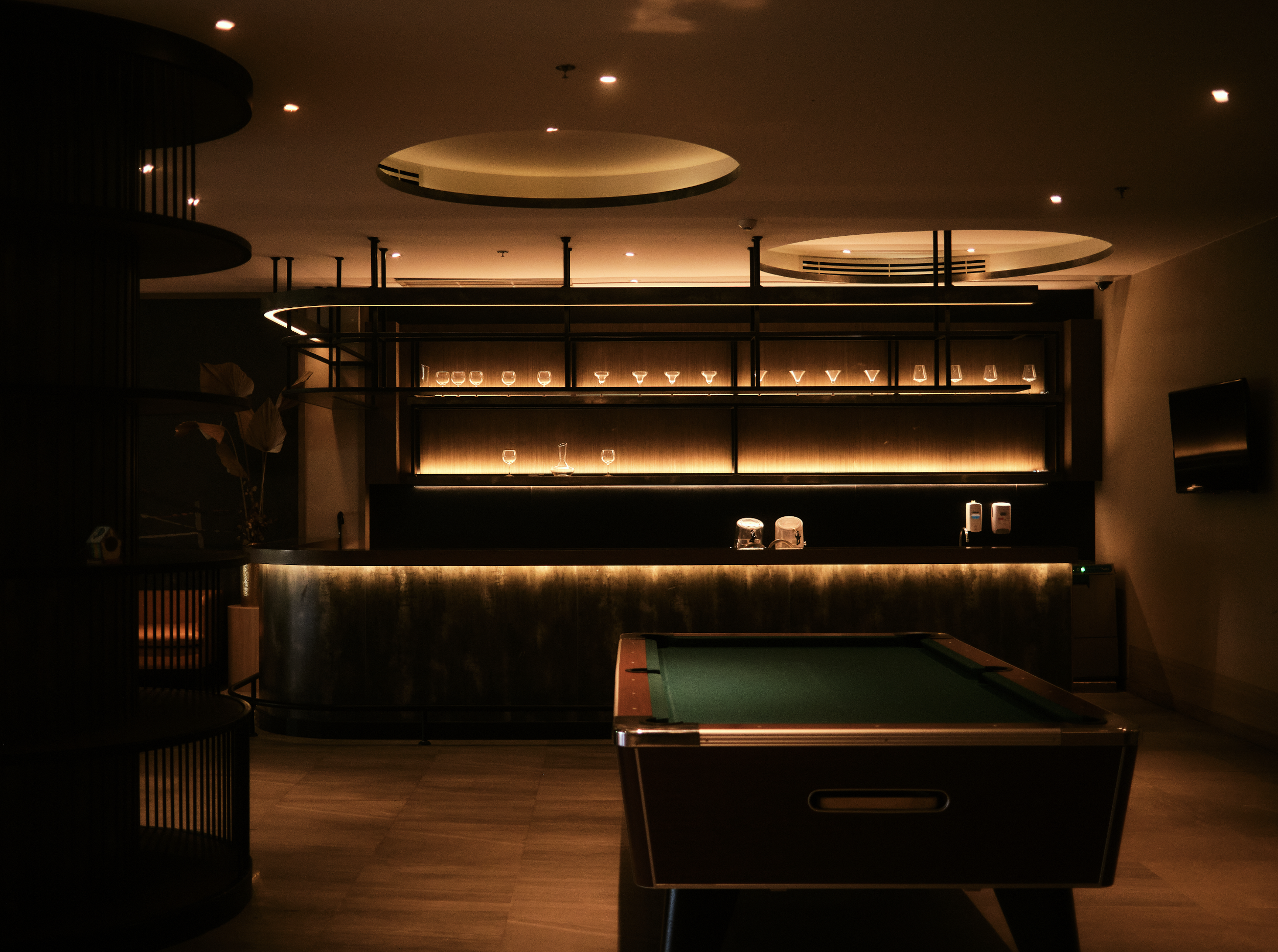 Dimly lit room with a bar area featuring glassware on shelves and a pool table in the foreground. Anemoia.