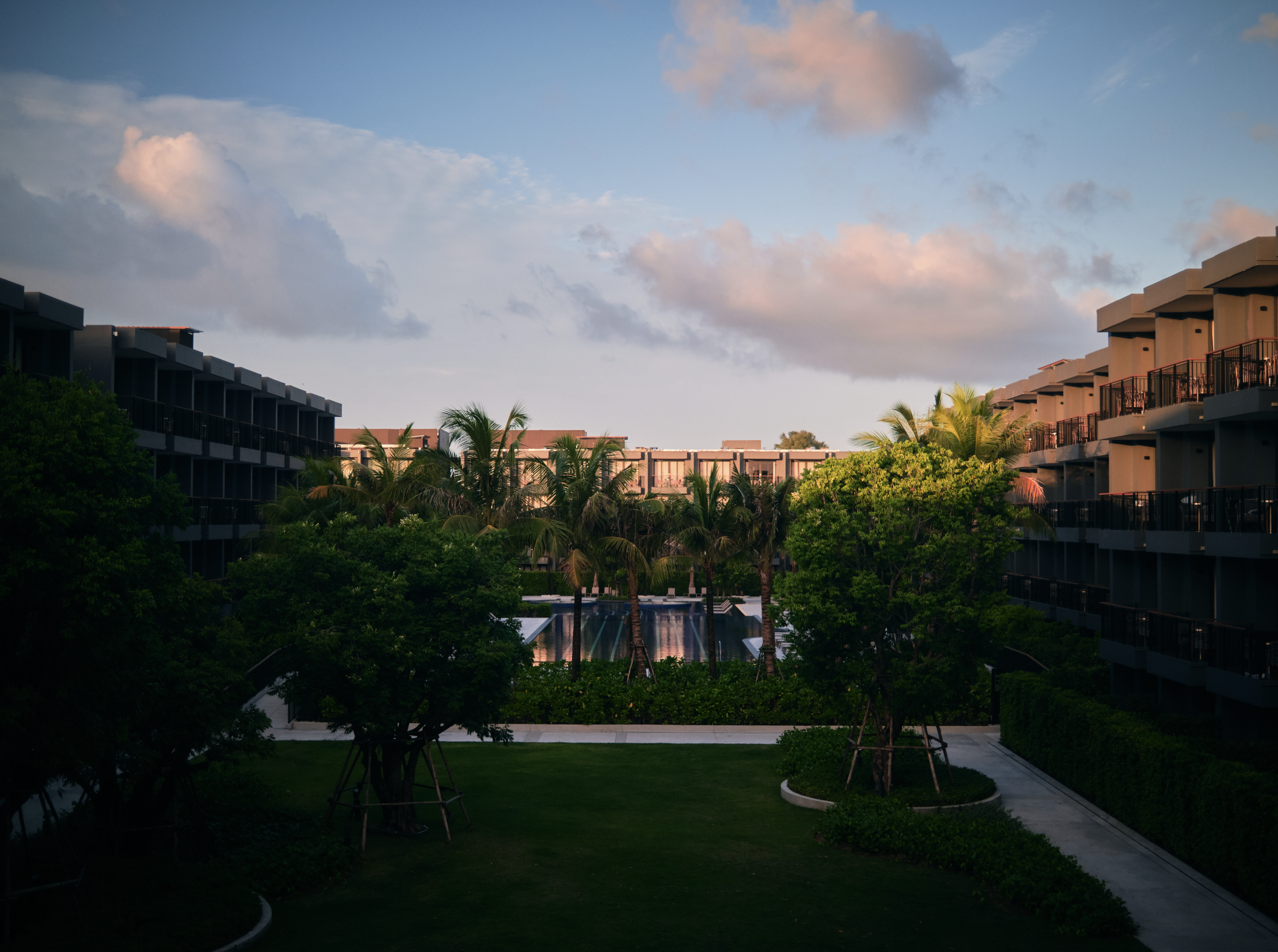 Residential apartment complex with multiple balconies, lush green trees, palm trees, and a clear sky with some clouds at sunset.