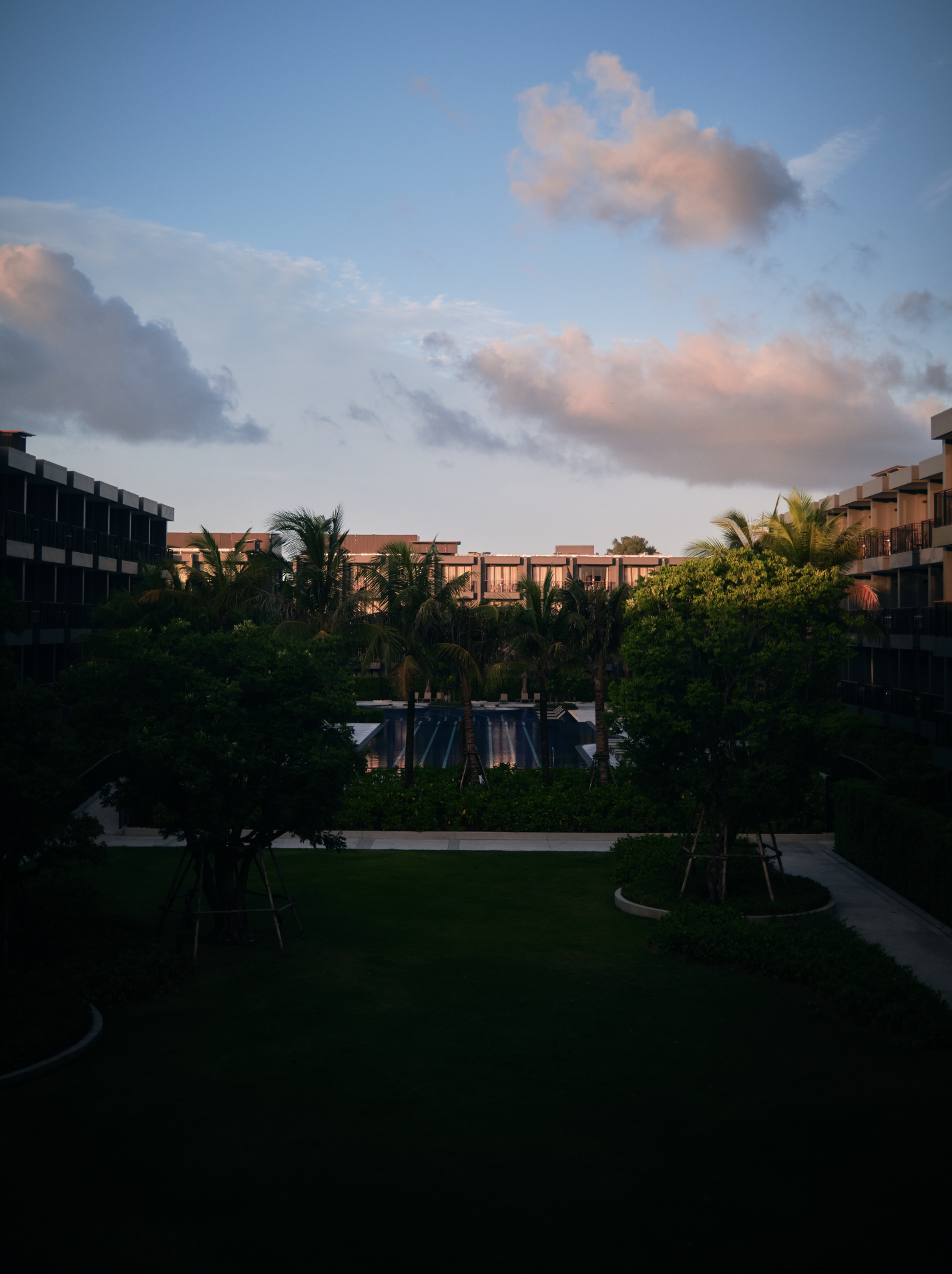 A view of an apartment complex with multiple buildings, surrounded by palm trees and greenery, during sunset with a partly cloudy sky.