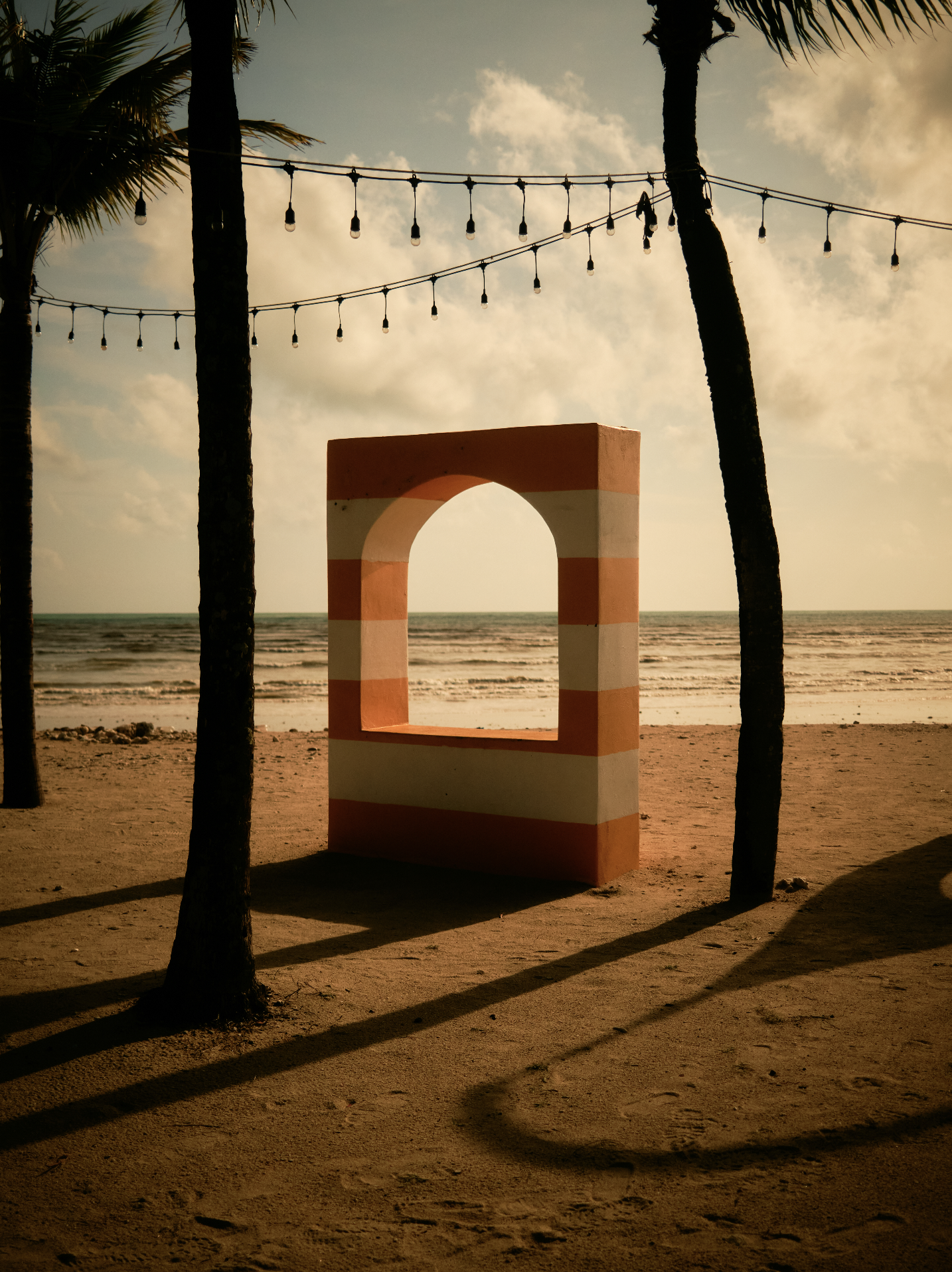 Beach scene at sunset with two tall palm trees, string lights hanging above, a striped pink and white archway, and the ocean in the background. Anemoia.