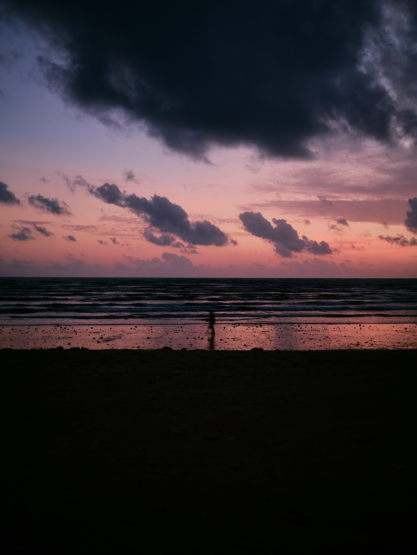 Sunset at the beach with dark clouds in sky and a person walking along the shoreline.