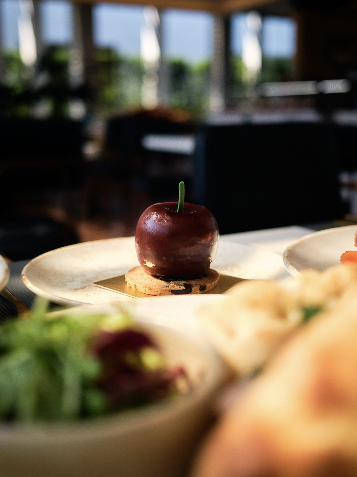 A shiny red apple-shaped dessert with a green stem on a cookie, placed on a white plate in a cozy indoor setting with blurred background of large windows and greenery.