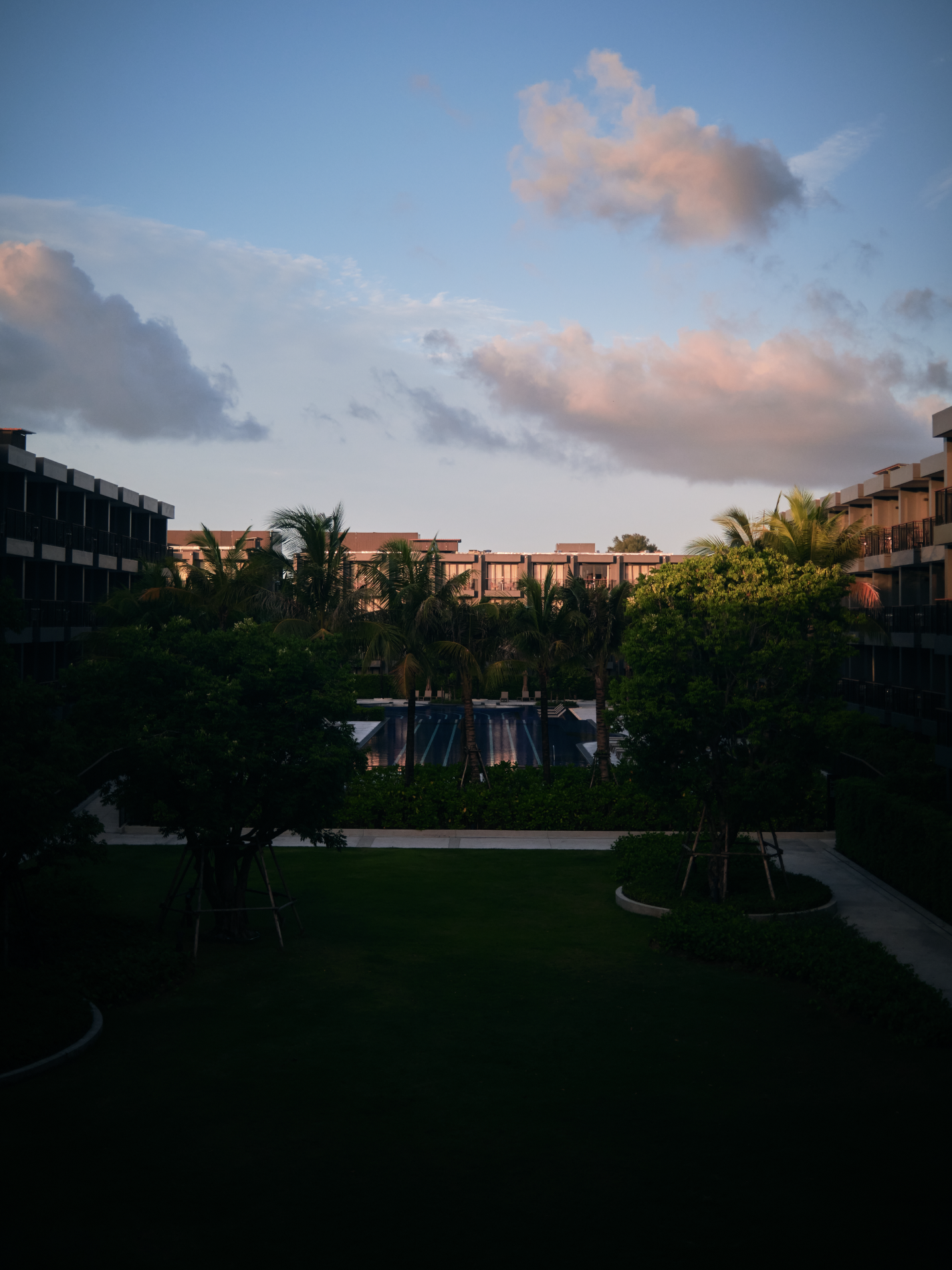 A view of a modern hotel or apartment complex with balconies, surrounded by lush green trees and palm trees, under a partly cloudy sky at sunset.