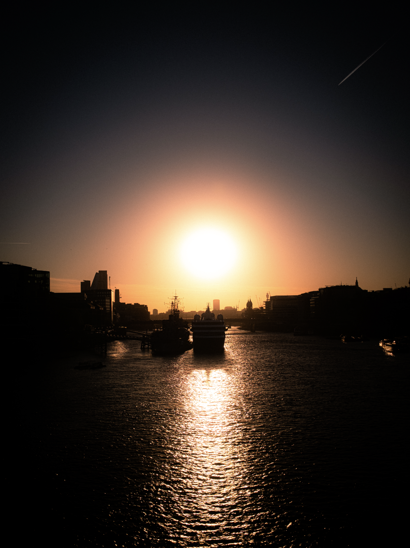 A city skyline silhouette at sunset with ships in the water and contrails in the sky.