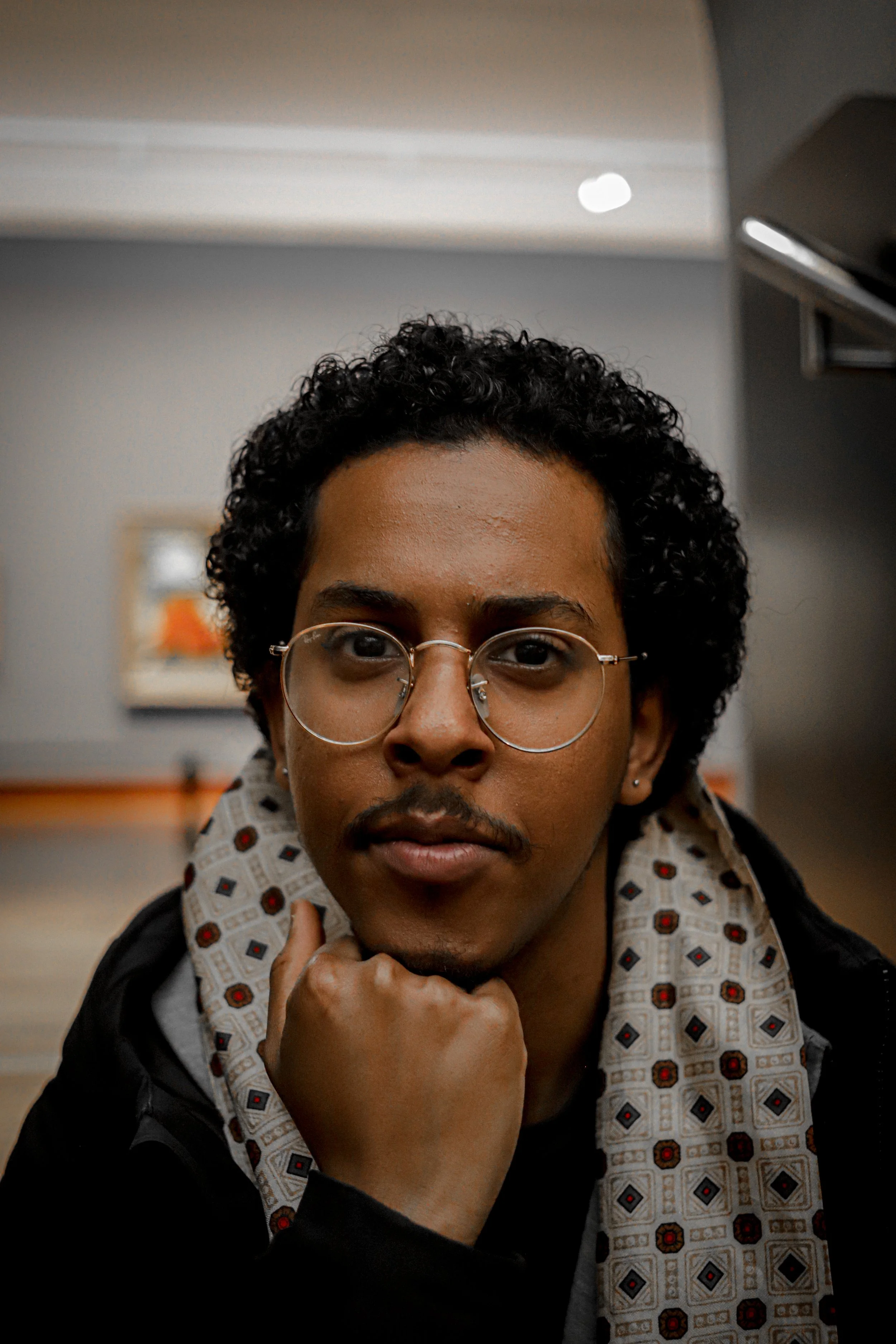 Close-up portrait of a young man with curly hair, wearing glasses, resting his chin on his hand, indoors.