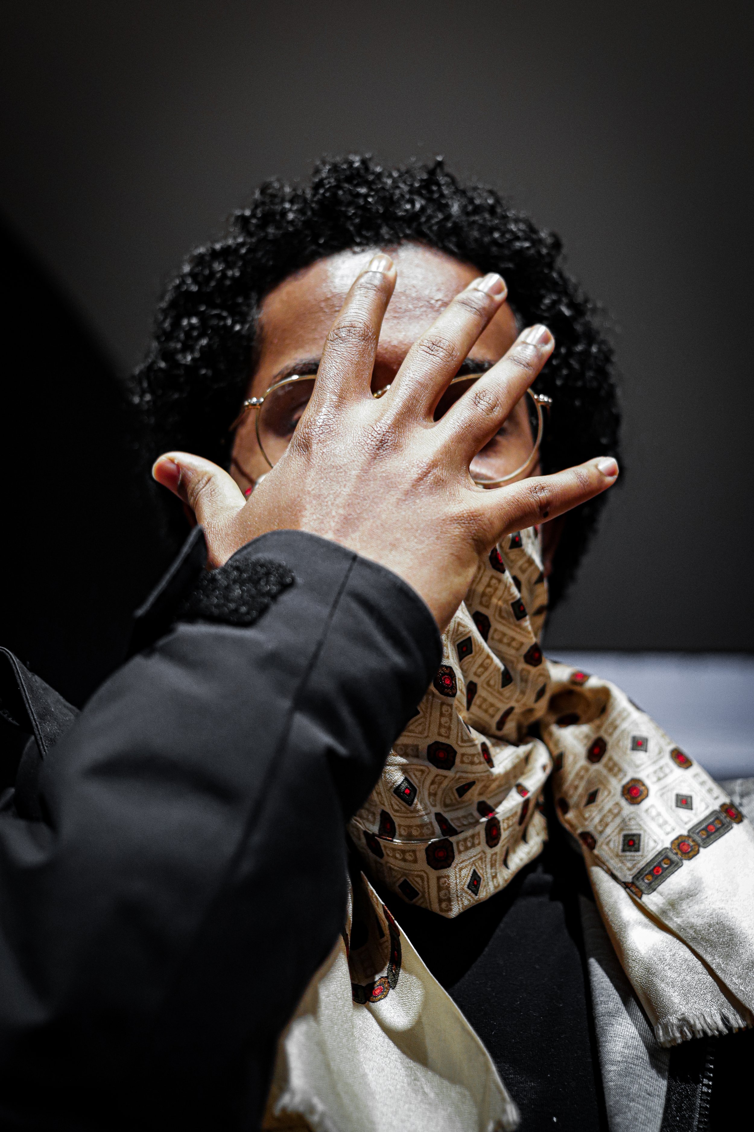 Person with curly hair and glasses, covering their face with their hand, wearing a patterned scarf and a black jacket.