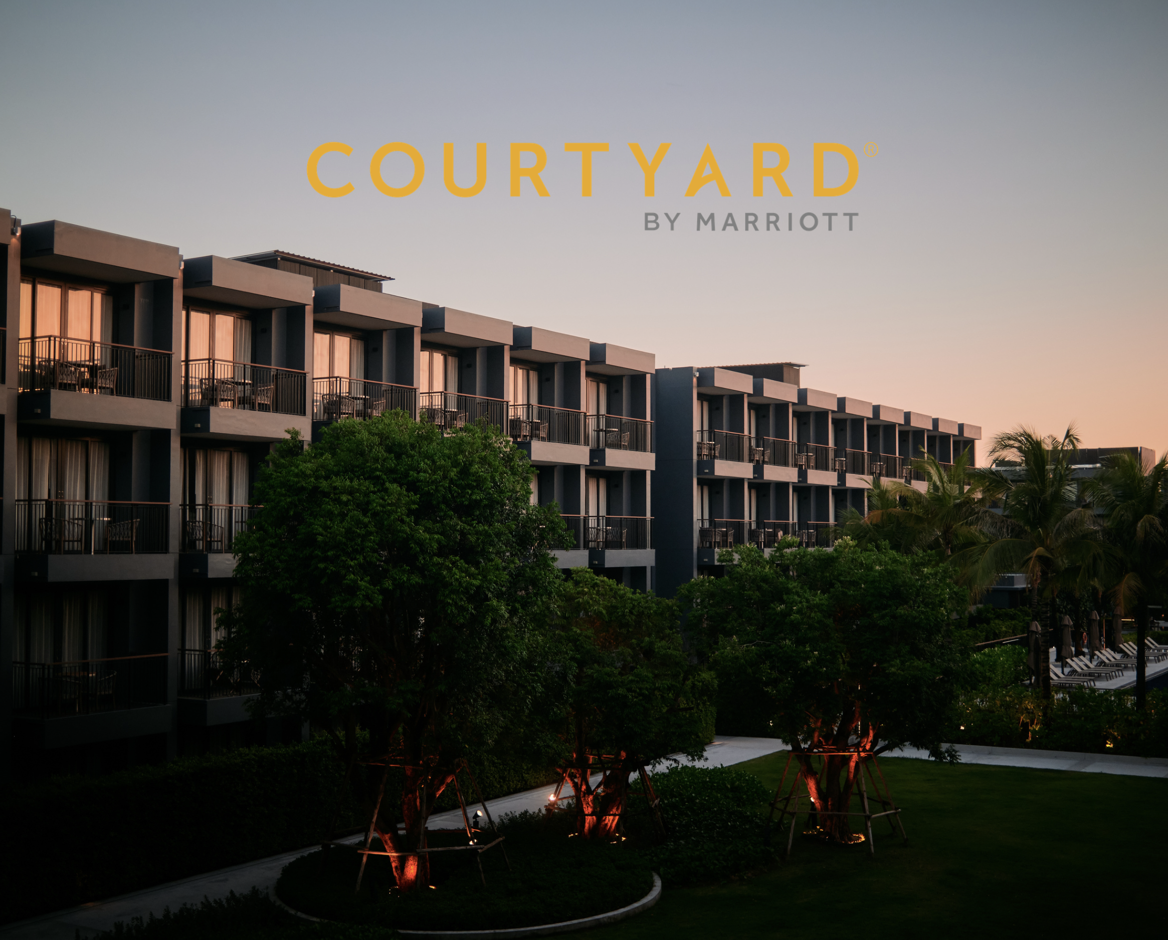 Exterior view of a modern hotel building with multiple balconies, surrounded by greenery and lit by outdoor lights at sunset.