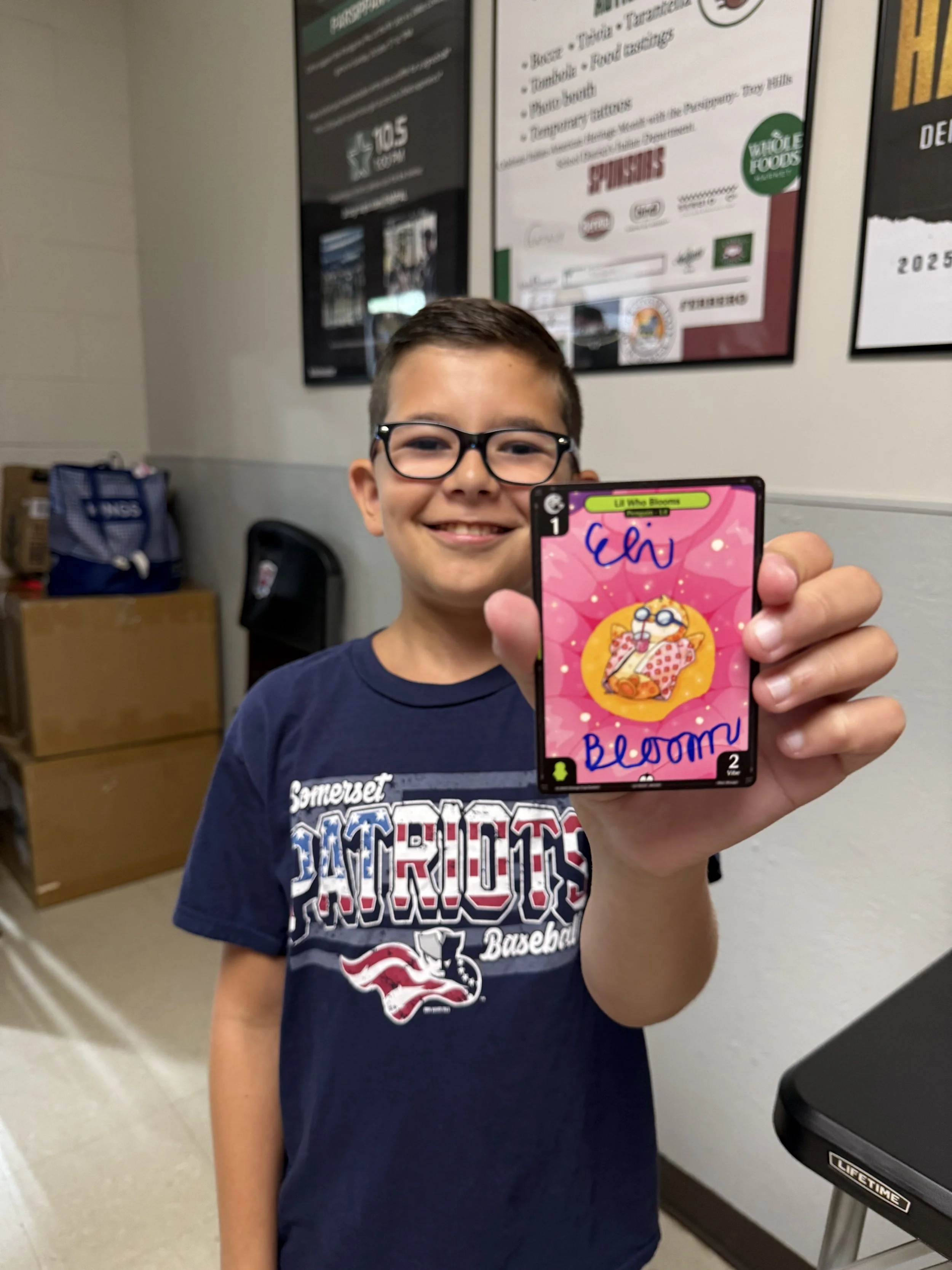 A young boy with glasses and a navy blue T-shirt smiling and holding up a card with an illustration of a taco and the words 'Ela Bloom' written on it.
