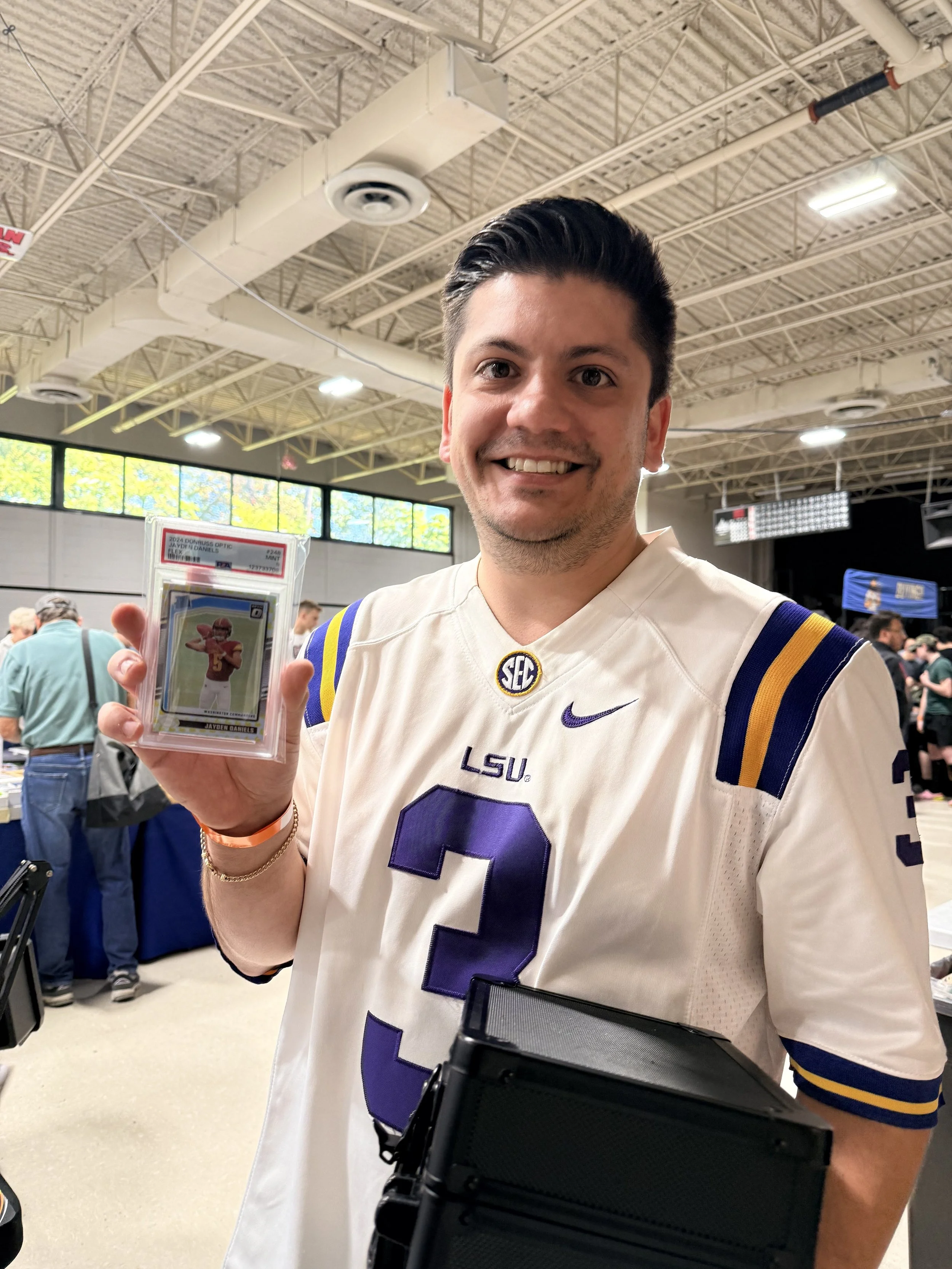 A man in a white LSU football jersey holding a trading card at a sports memorabilia fan event.