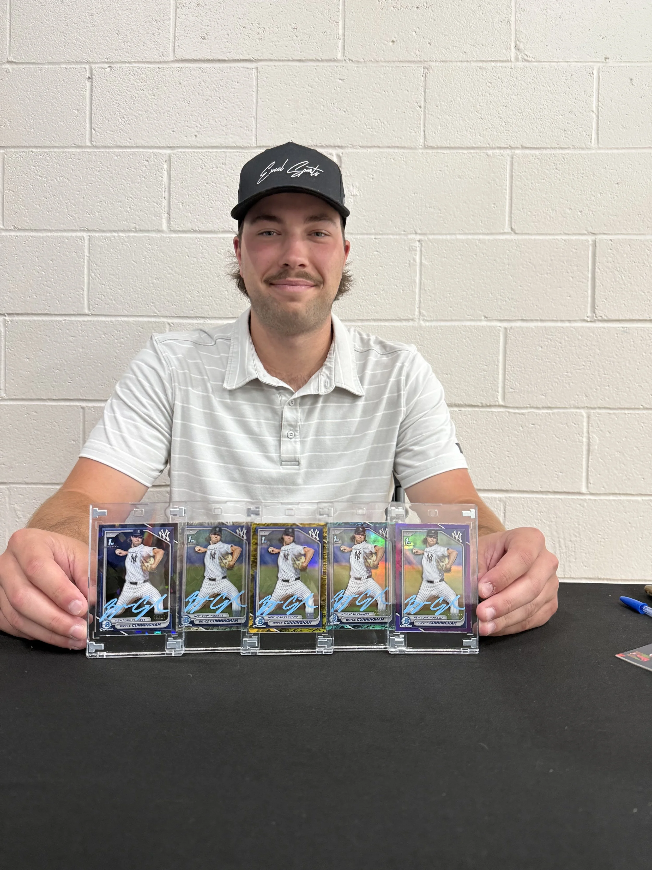 A man wearing a black cap and a white striped polo shirt sitting at a table with five collectible baseball cards of a player in a New York Yankees uniform, encased in plastic display cases.