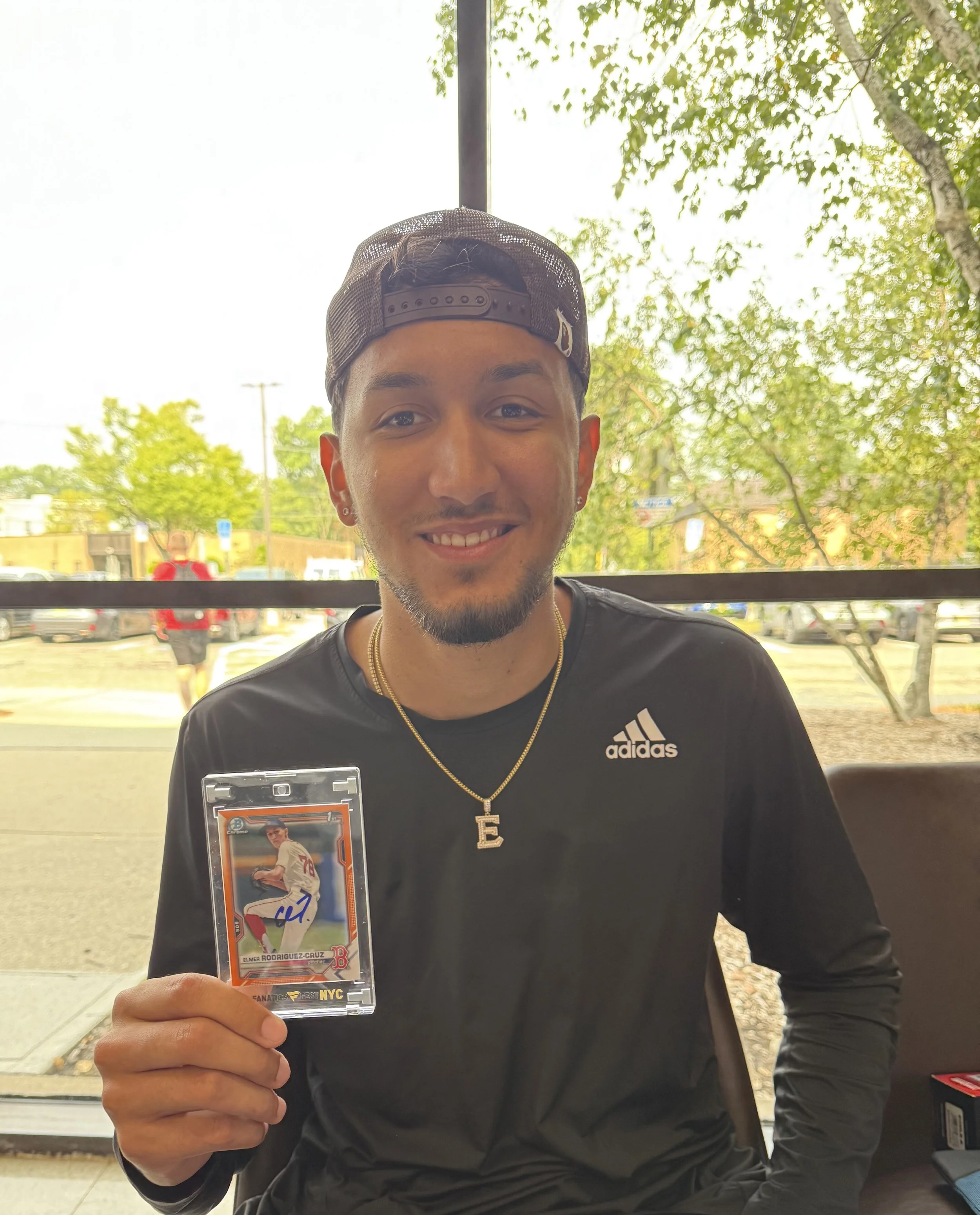 Young man with a beard and mustache wearing a black Adidas shirt and a black cap backwards, holding a baseball trading card inside a clear plastic sleeve, smiling at the camera.