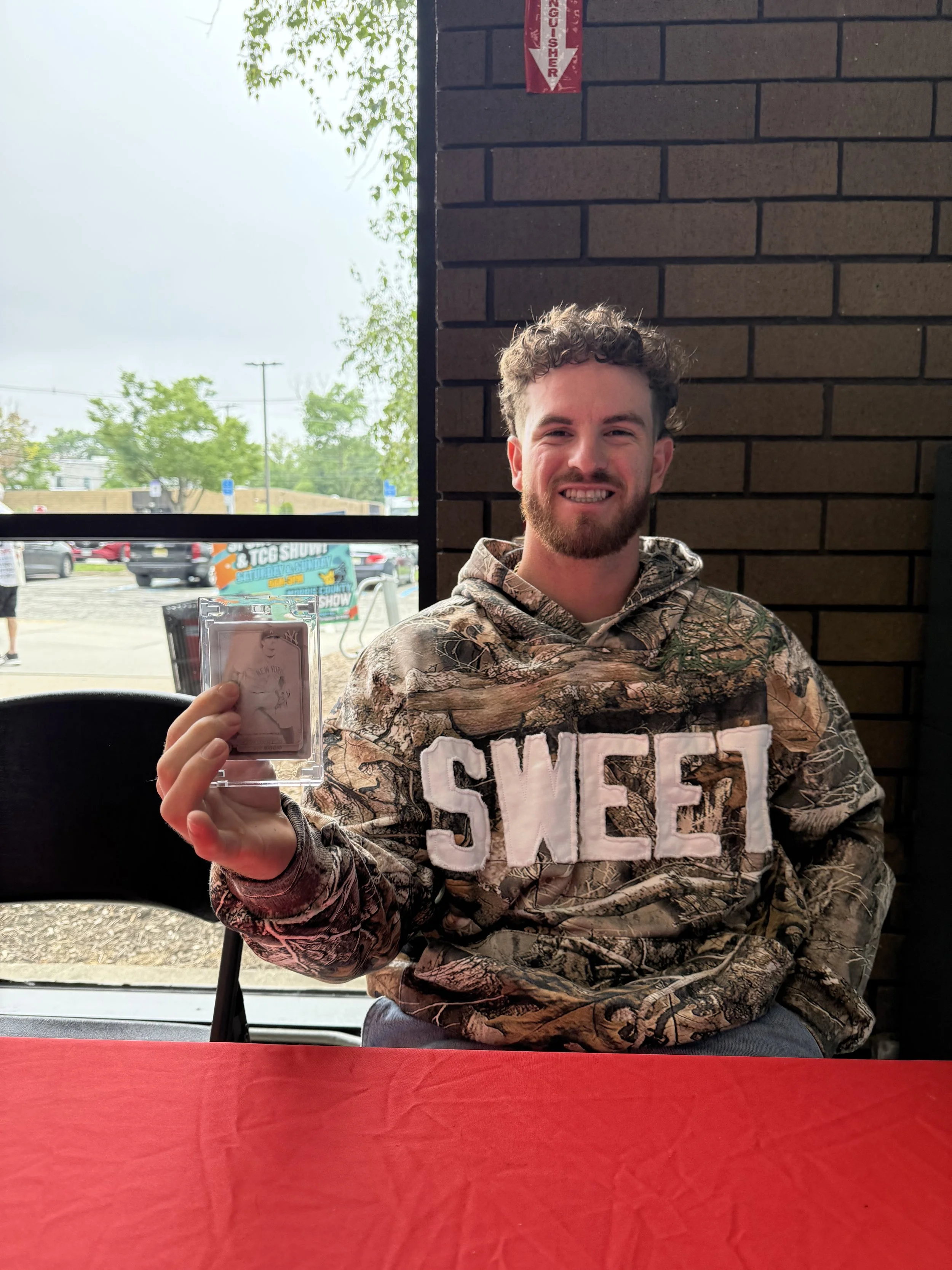 A man with curly hair and a beard smiling, wearing a camouflage hoodie with the word 'SWEET' in large white letters, sitting at a red table, holding a trading card in a plastic case, inside a restaurant with a brick wall and window behind him.