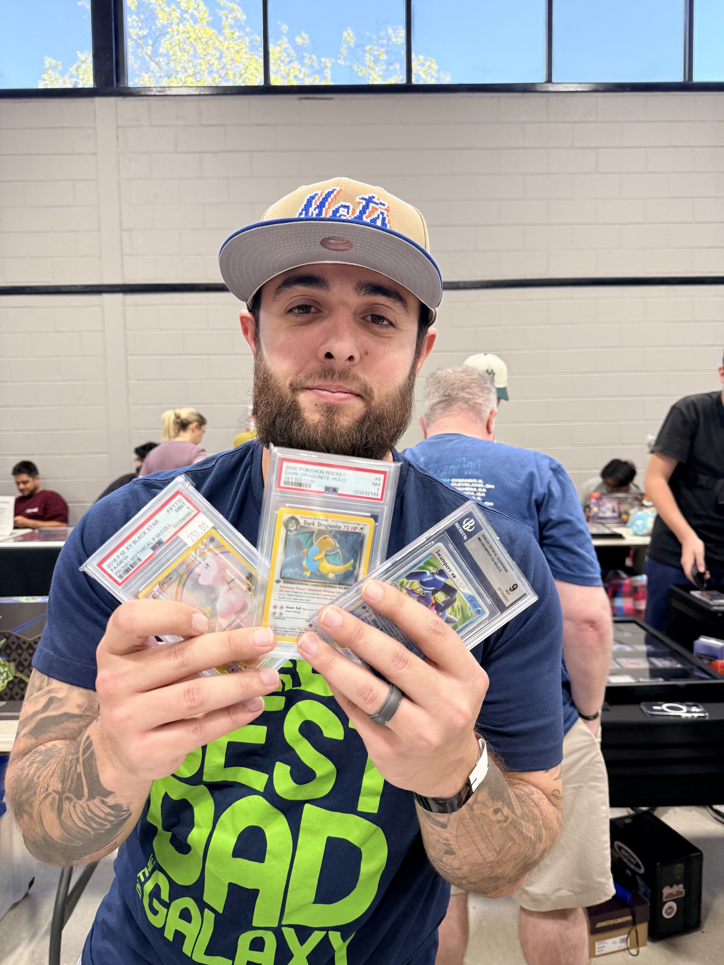 A man with a beard, wearing a Mets baseball cap and a navy blue T-shirt that says "The Best Dad in the Galaxy," showing four Pokémon cards at a table in a large indoor space, possibly at a trading card event.