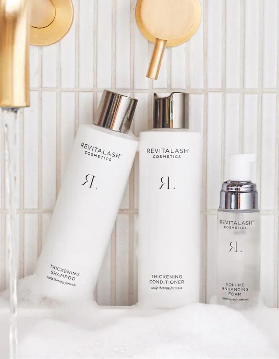 Three Revitalash Cosmetics hair care bottles in a shower with white tiled wall background. The bottles include thickening shampoo, thickening conditioner, and volume enhancing foam.