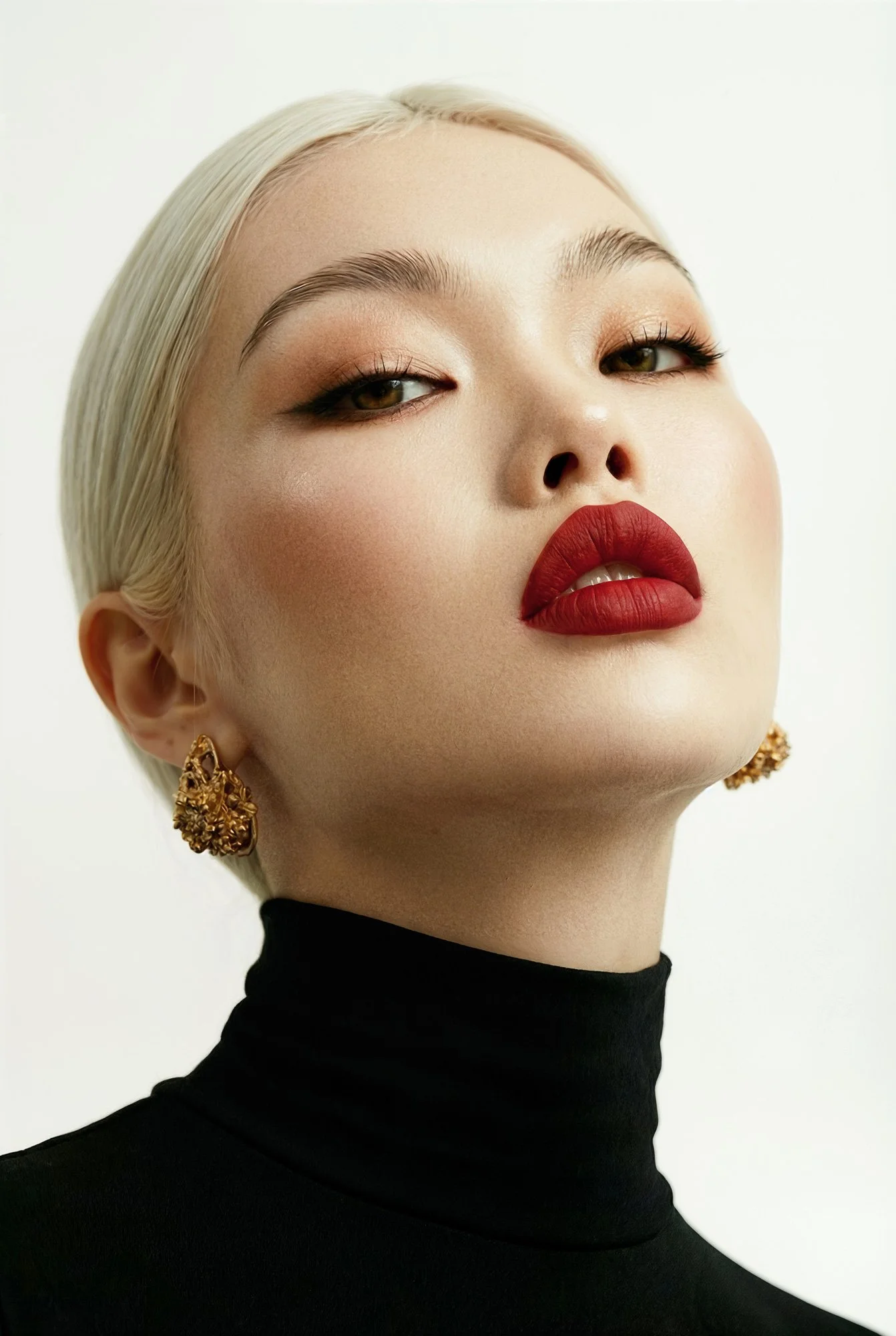 Low-angle beauty portrait of a woman with platinum blonde hair and red lipstick, wearing a black turtleneck and gold earrings, with natural skin texture and soft studio lighting.