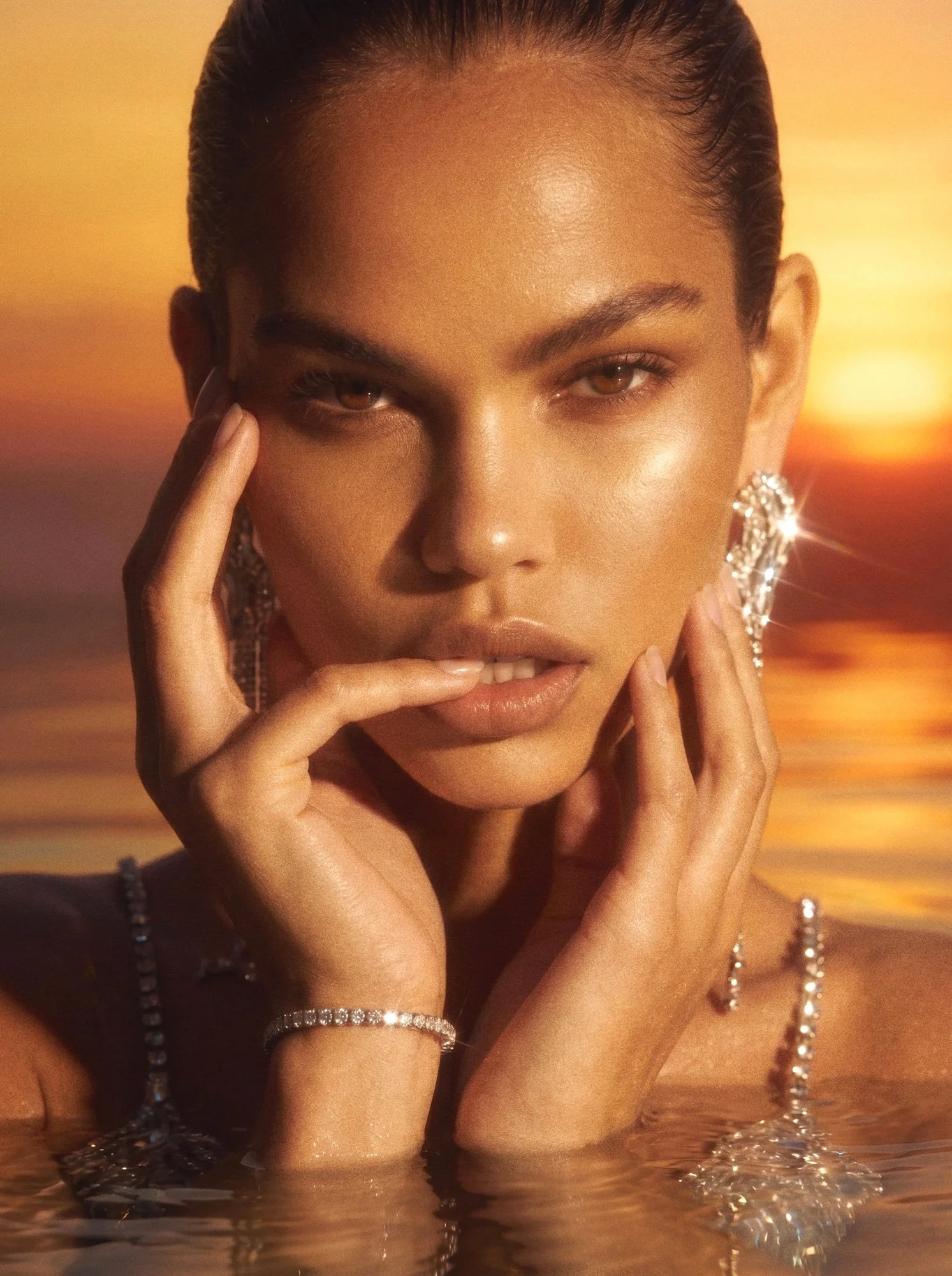 Female model in reflective water at sunset wearing a sparkling luxury gown and statement jewellery, captured in golden cinematic light. By Yannick De Bakongo