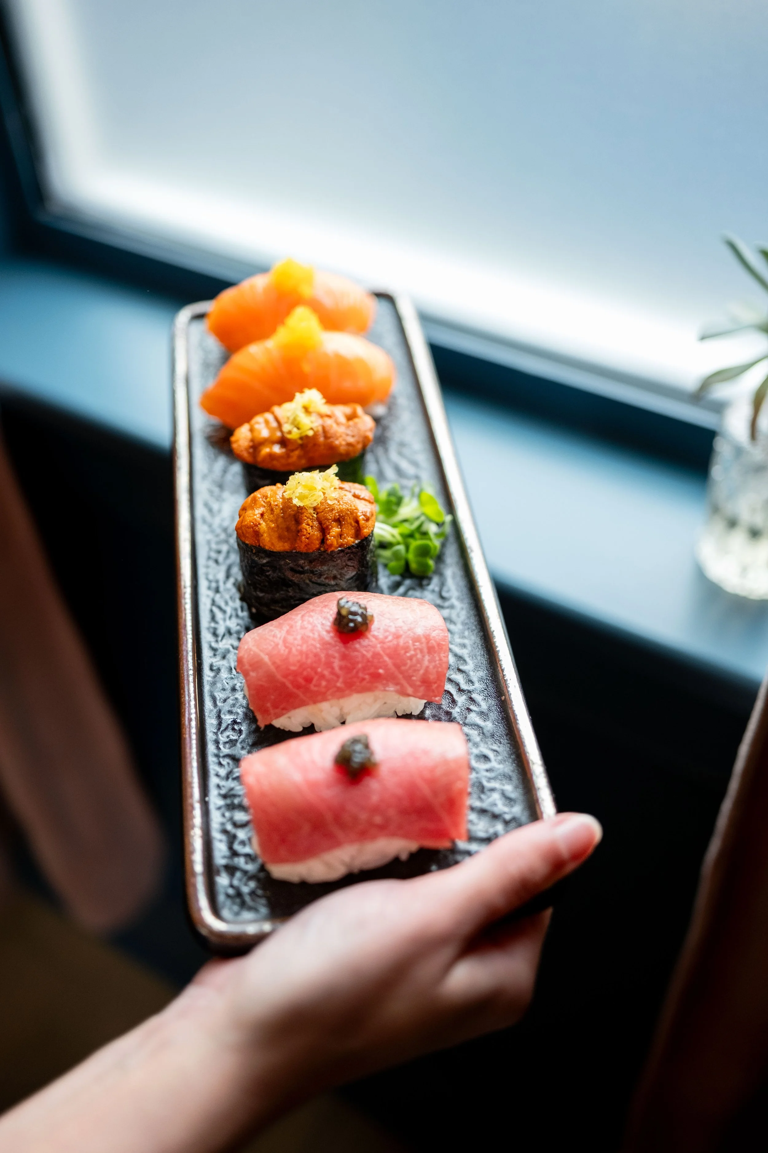 Hand holding a platter of sushi rolls and sashimi including salmon and tuna, photographed near a window at Nichinan.