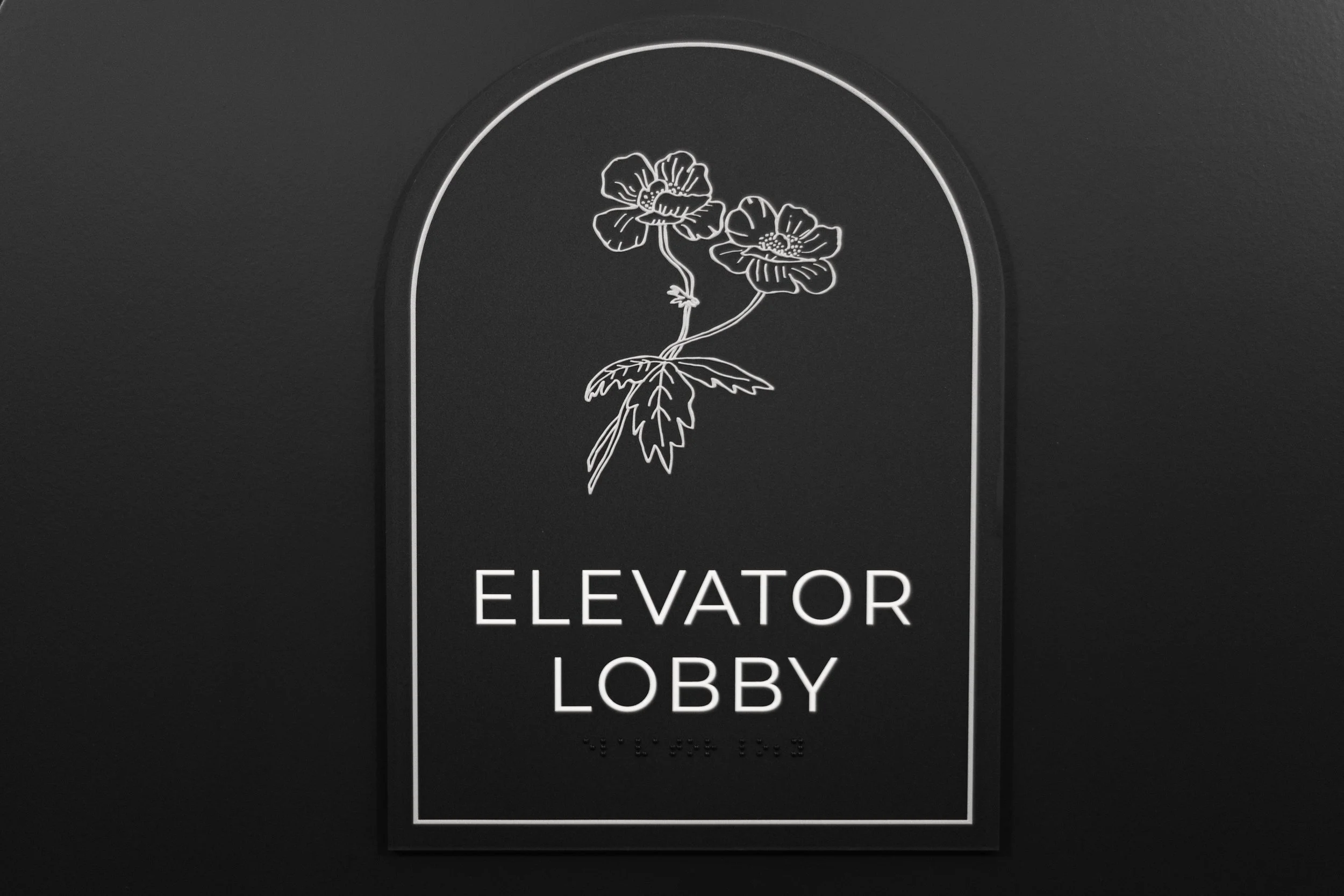 Black sign with white outline of two flowers and the text 'Elevator Lobby' underneath.