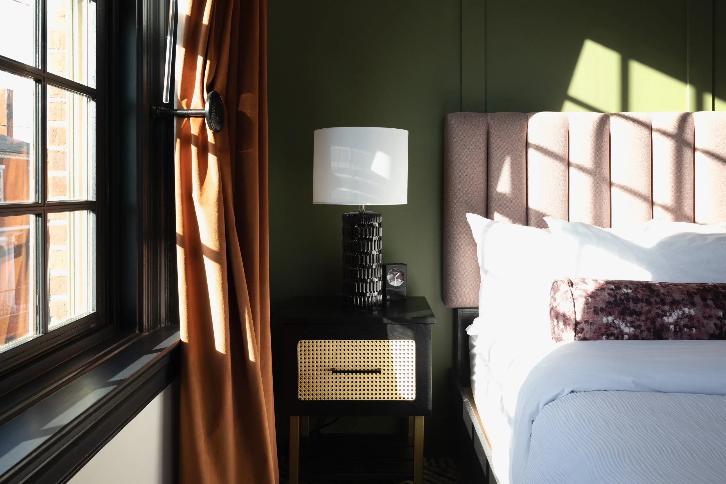 Sunlit bedroom corner with a window, brown curtains, a bedside table with a black textured lamp, a small radio, and a bed with white sheets, a pink pillow, and a tall beige headboard. Shadows cast across the wall and bed.