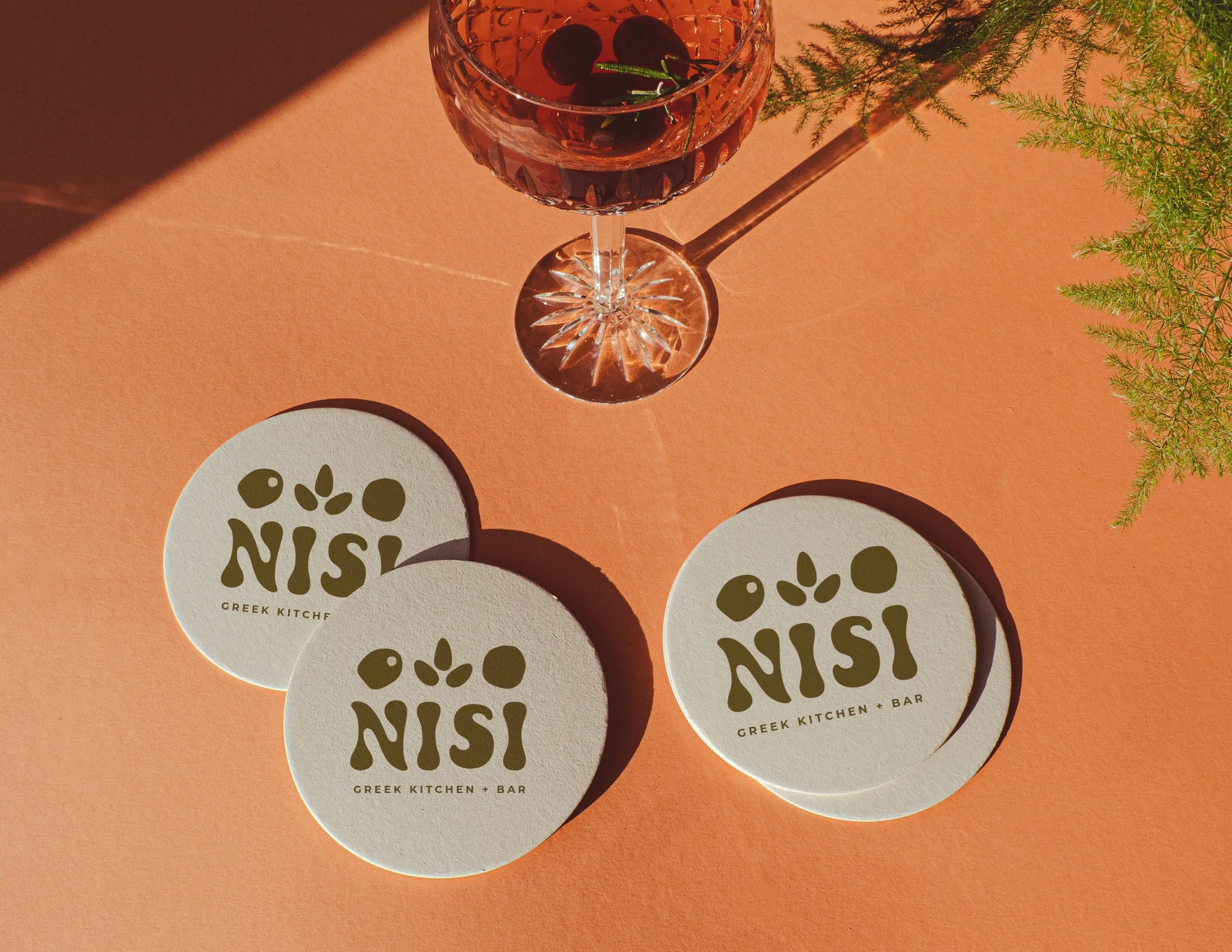 Branded Nisi Greek Kitchen + Bar coasters styled on a peach surface with a glass of red cocktail garnished with olives and herbs.