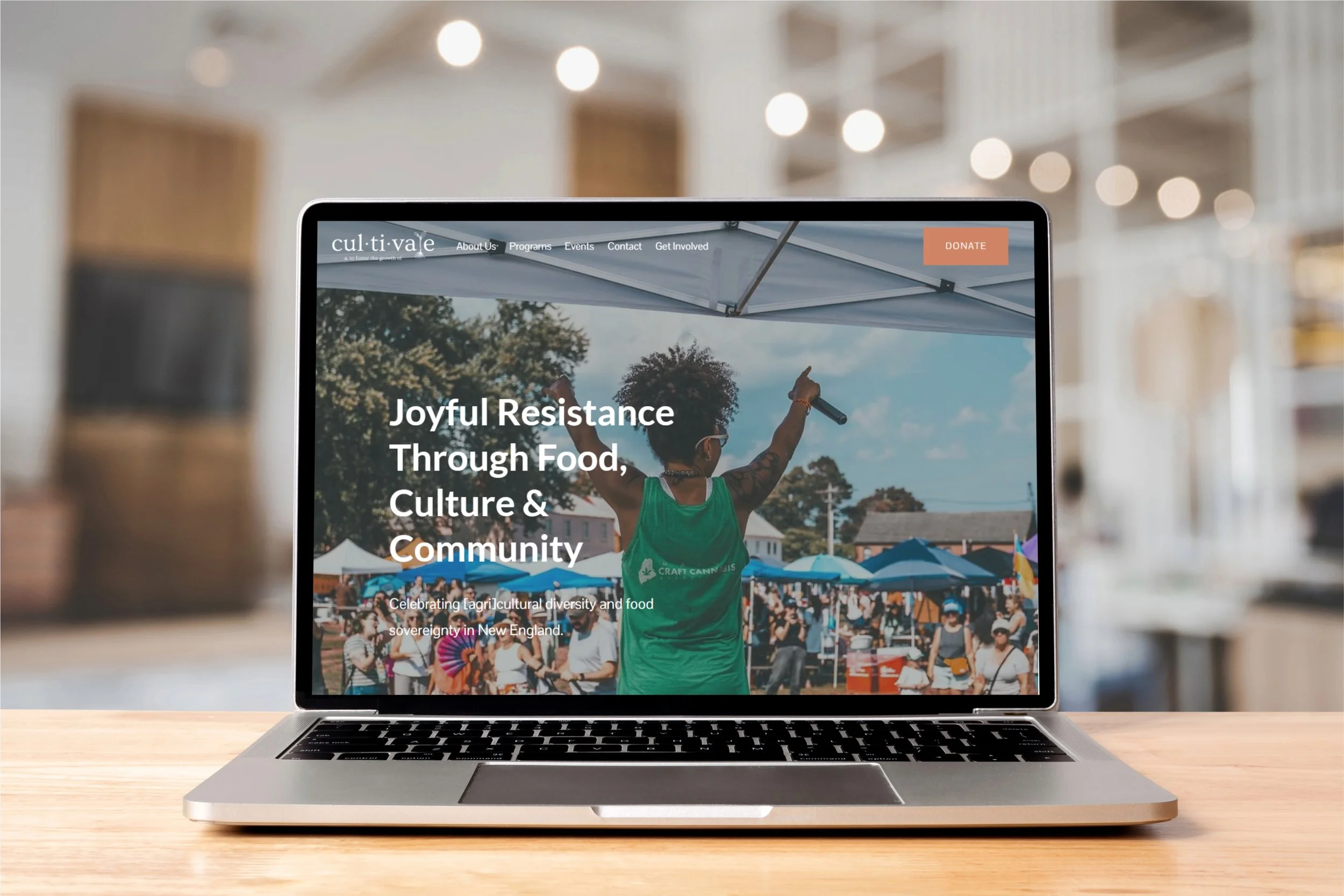 Laptop displaying a community event website with an image of a person speaking at an outdoor festival or gathering and the text 'Joyful Resistance Through Food, Culture & Community.'