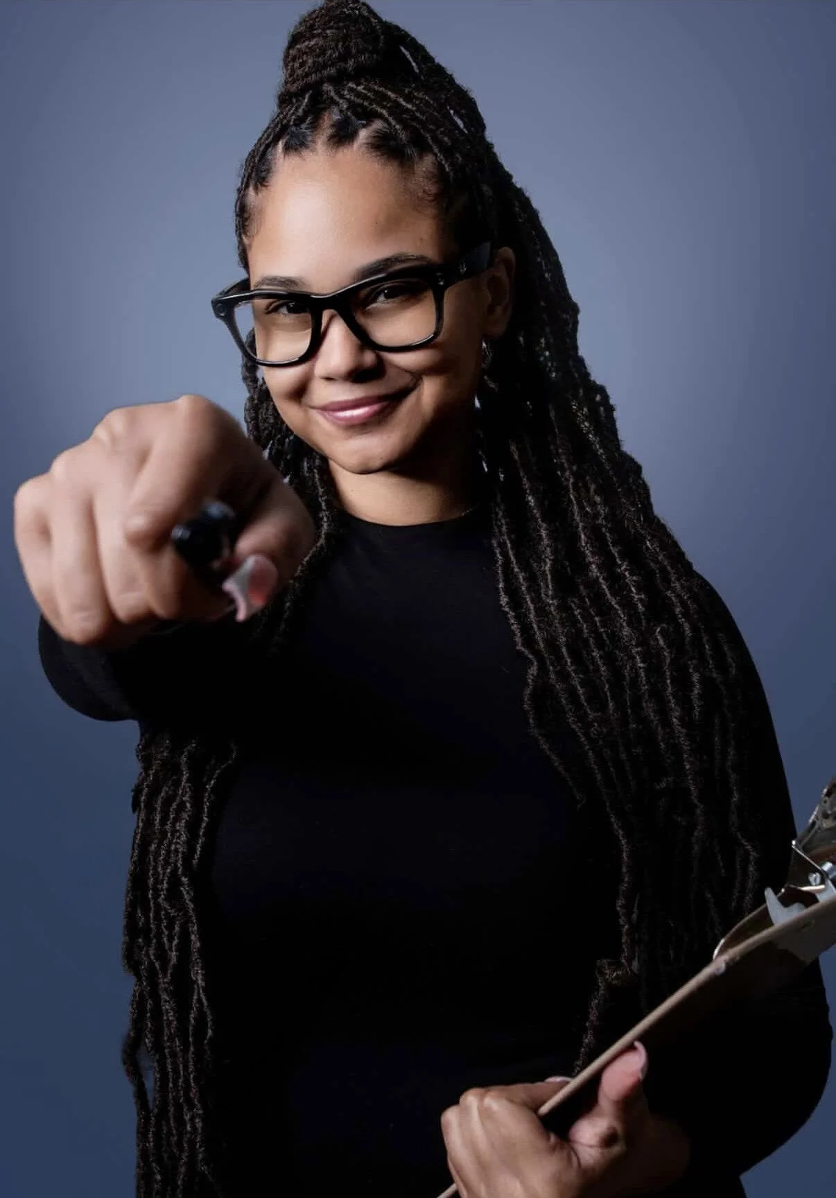 Dee, personal and executive assistant holds clipboard in one hand while pointing at the camera with a pen in her hand. She is wearing meta glasses and has locs in her hair.