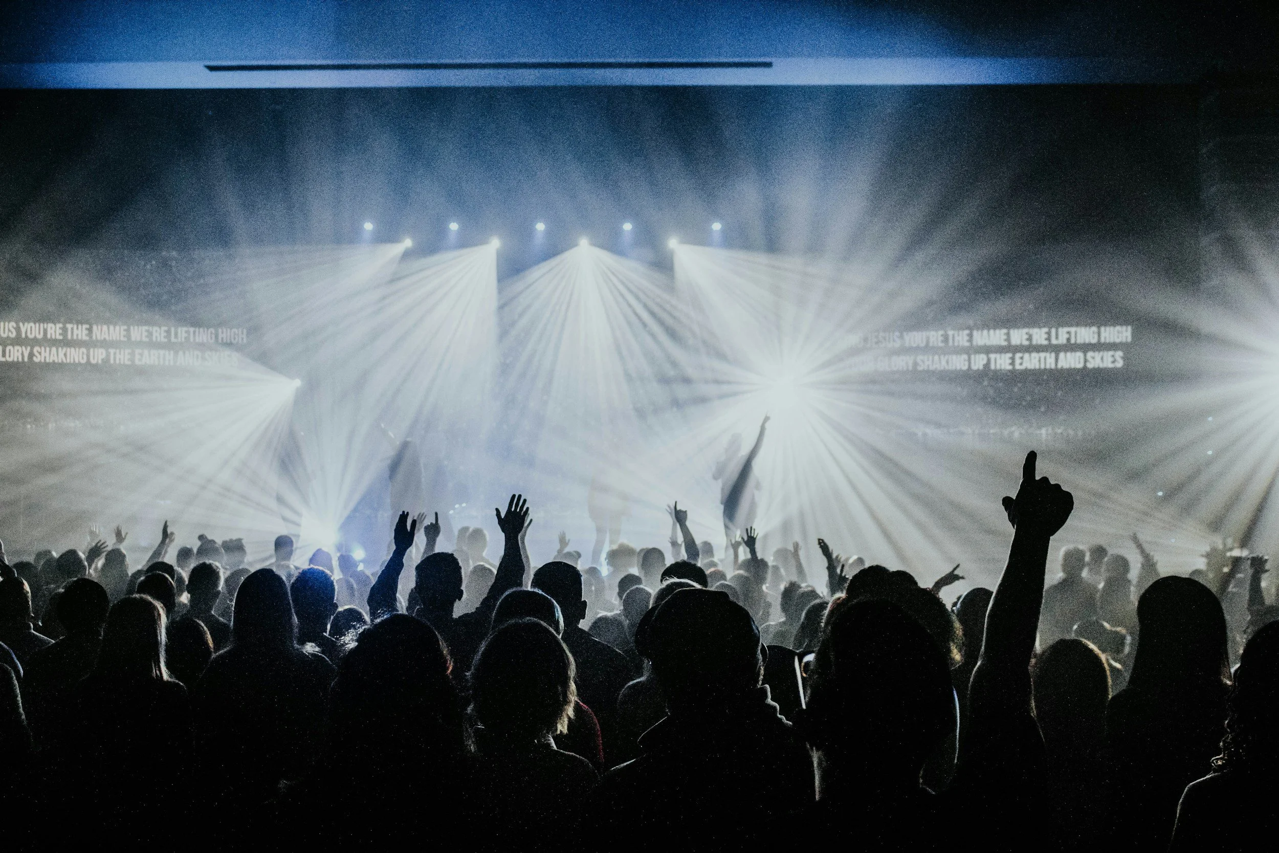 A crowd at a concert or event with hands raised, facing a stage illuminated with bright spotlights and light beams, creating an energetic atmosphere.