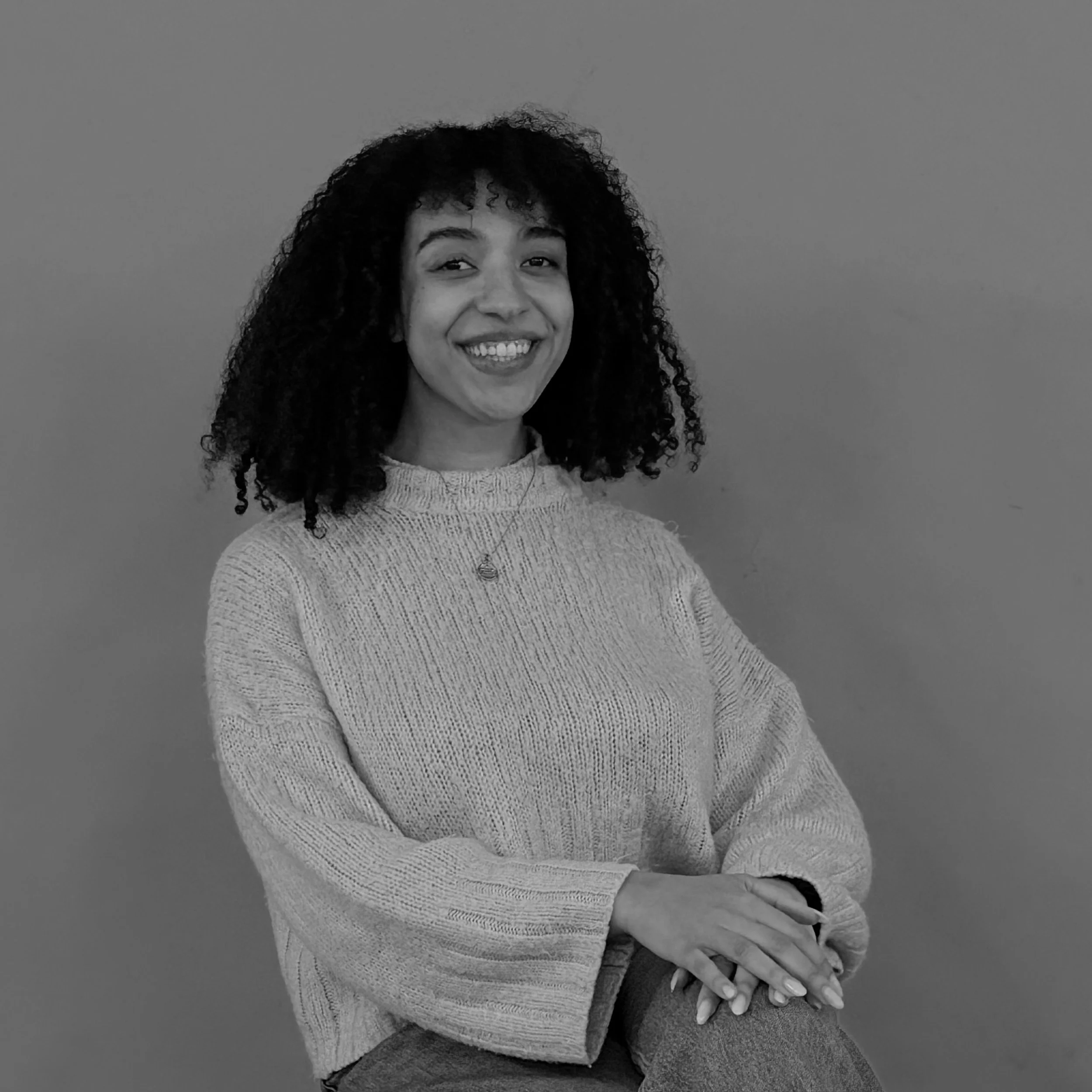 A woman with curly hair smiling, wearing a sweater, standing against a plain background.