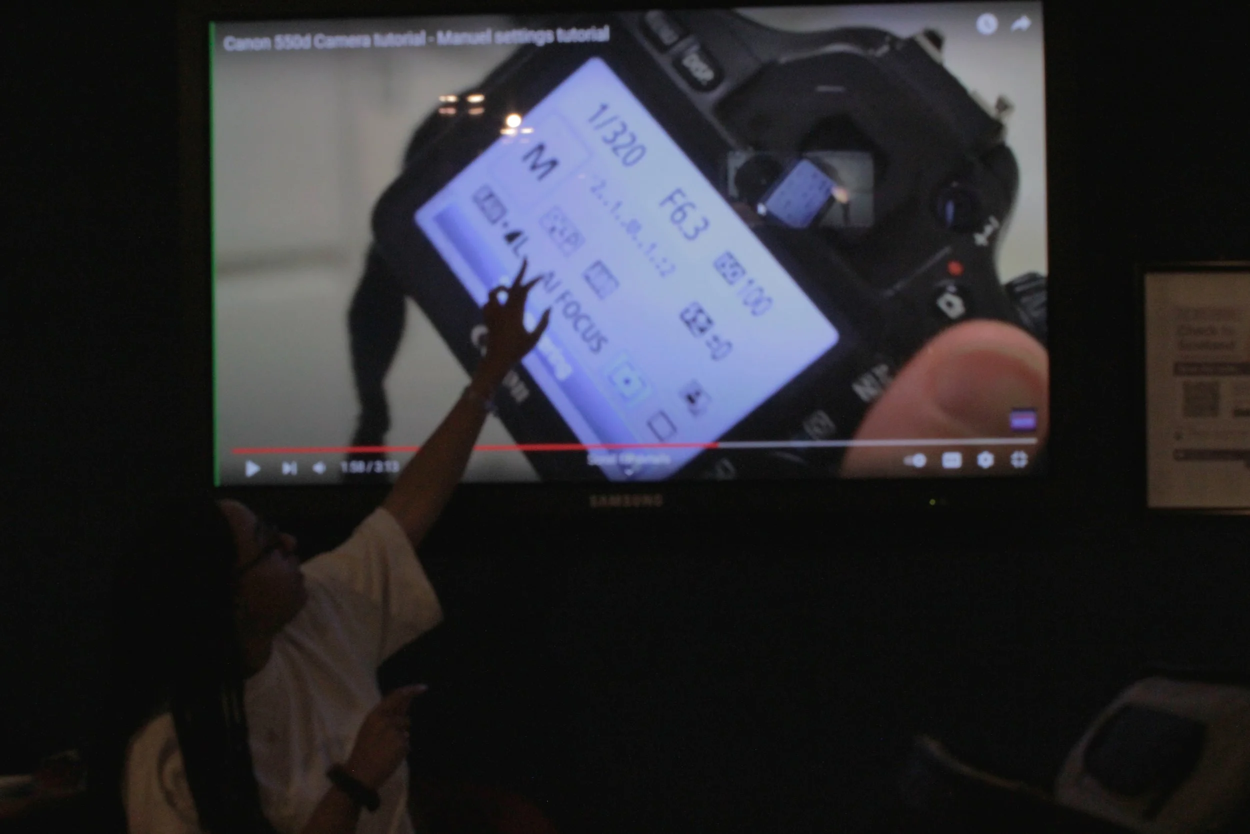 A person with a small hand reaches towards a camera's LCD screen on a Canon DSLR camera, which displays various settings and options. The scene is dark, with a television or monitor in the background showing a paused video.
