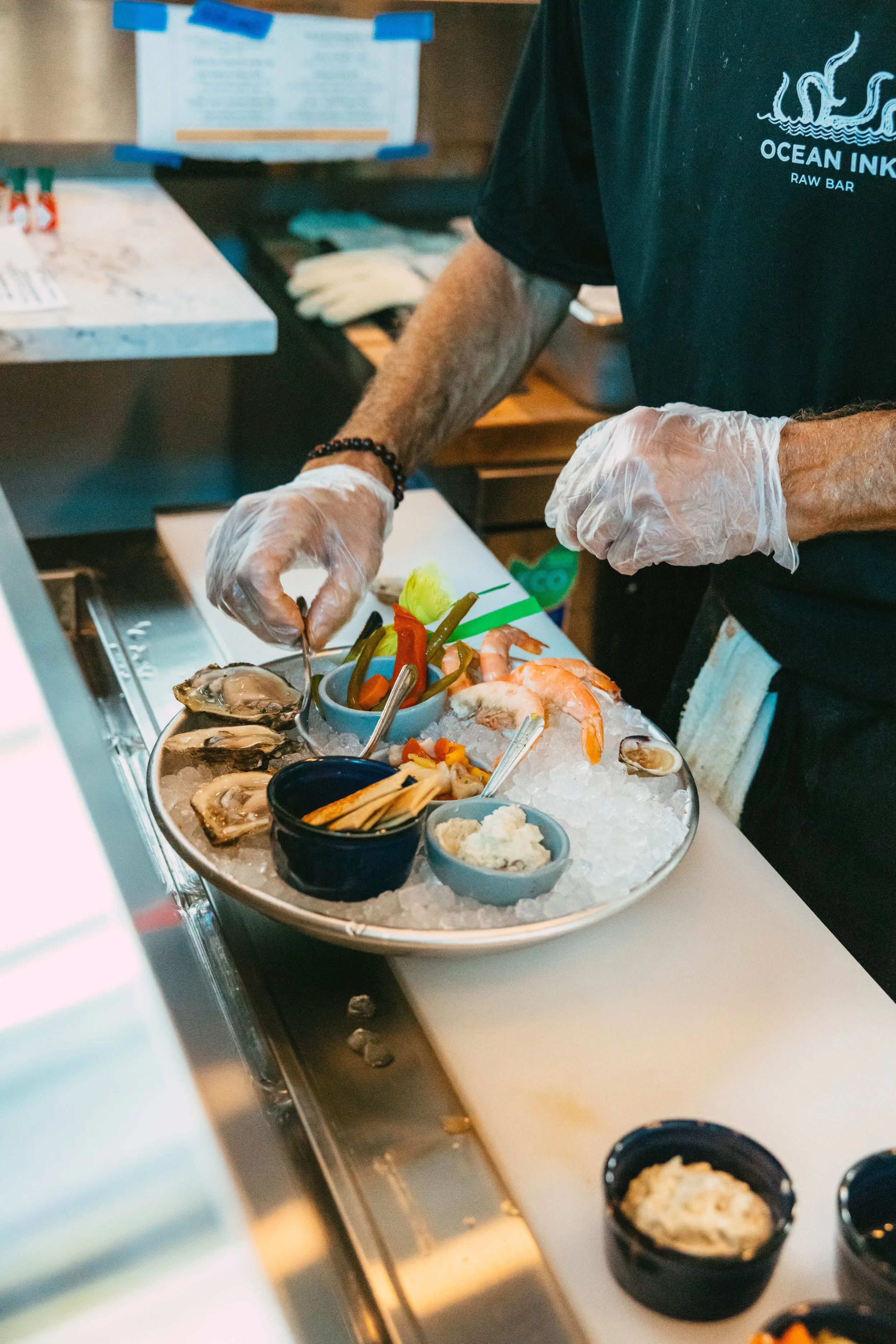 A person wearing a black Ocean Ink T-shirt and plastic gloves preparing a seafood platter with oysters, shrimp, and various dipping sauces on ice.