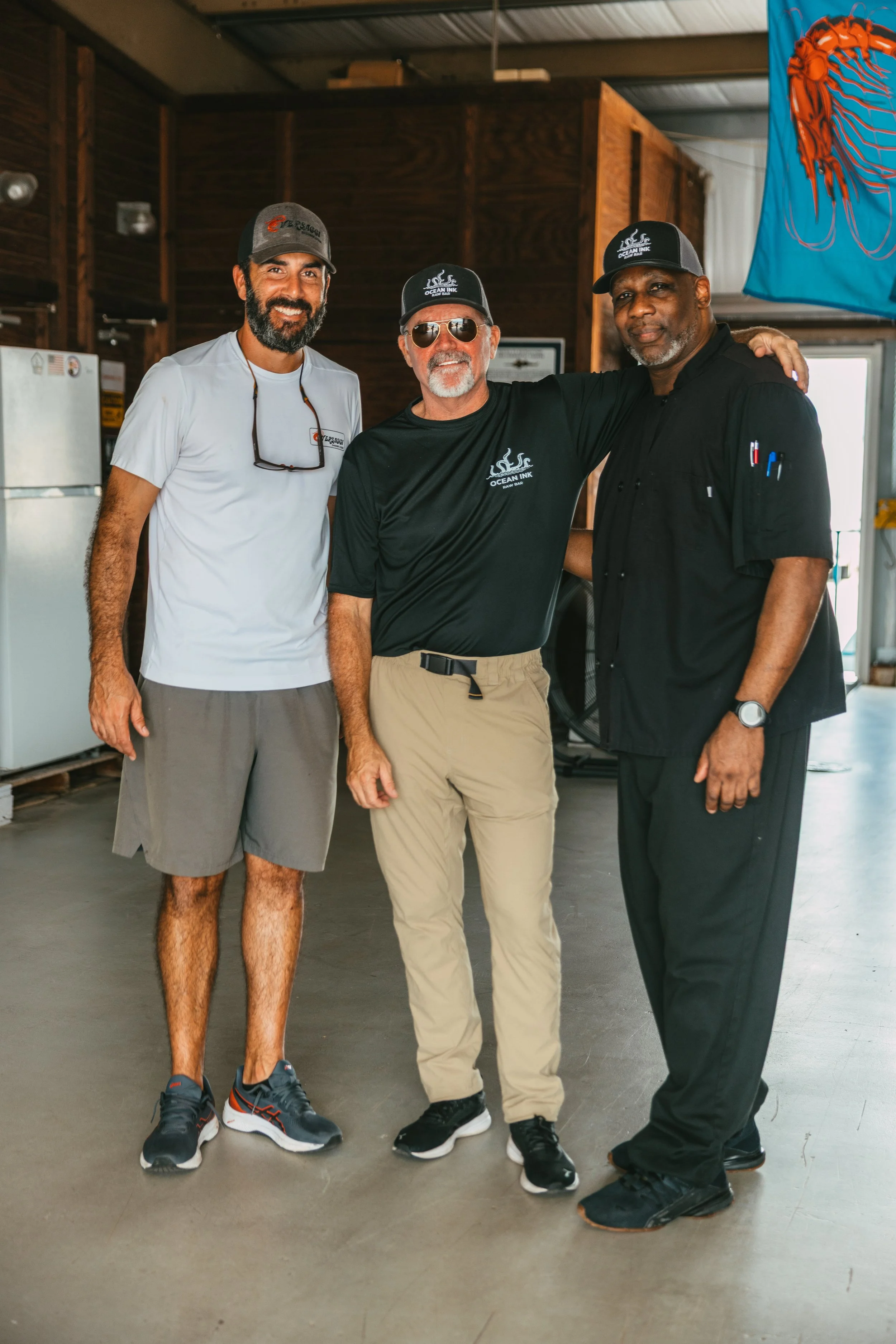 Three men standing inside a wooden indoor space with a large jellyfish banner on the wall behind them. They are smiling, with the middle man having his arms around the others.