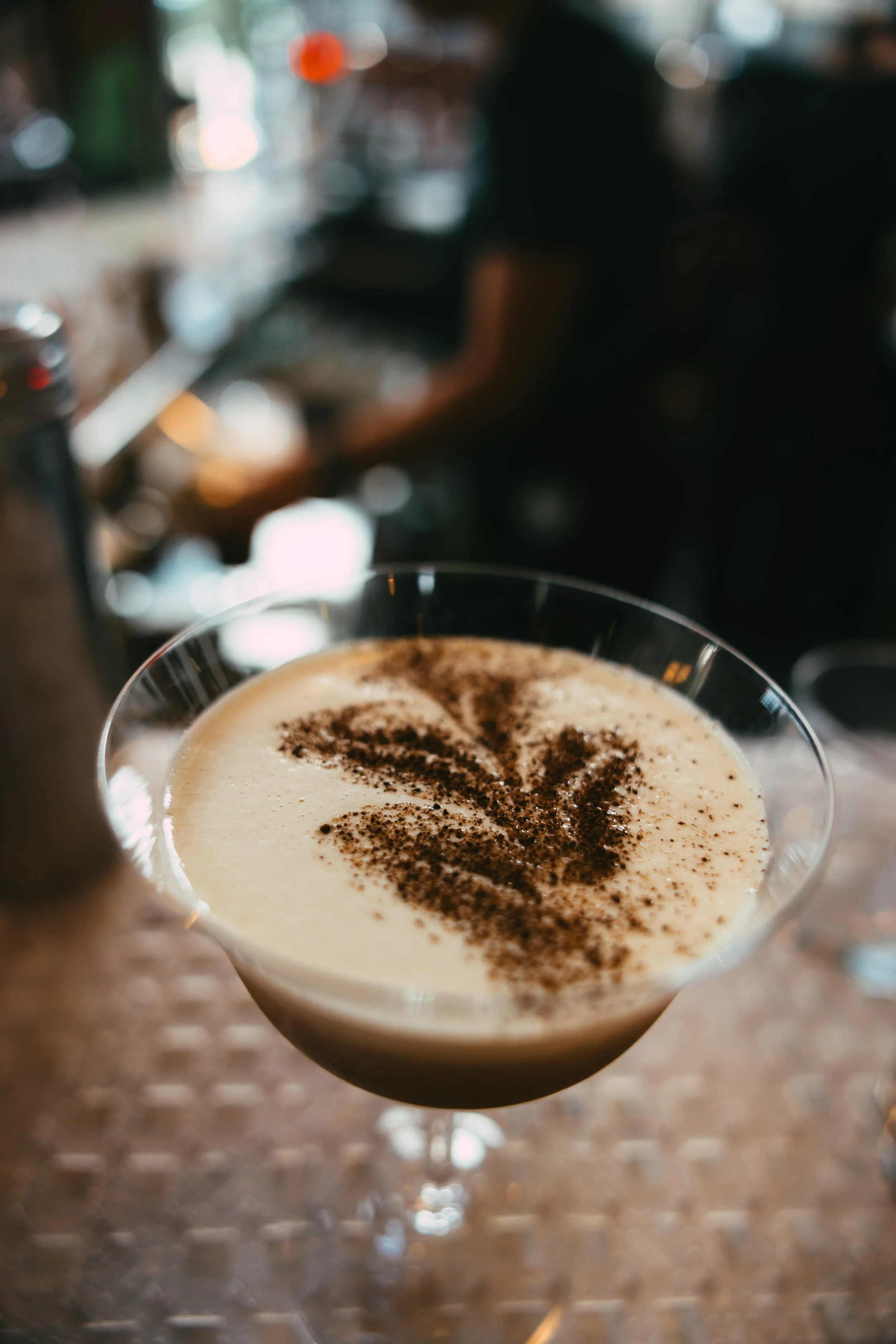 A close-up of a cocktail glass filled with a creamy drink topped with coffee or cocoa powder in a leaf design, on a bar counter in a dimly lit setting.