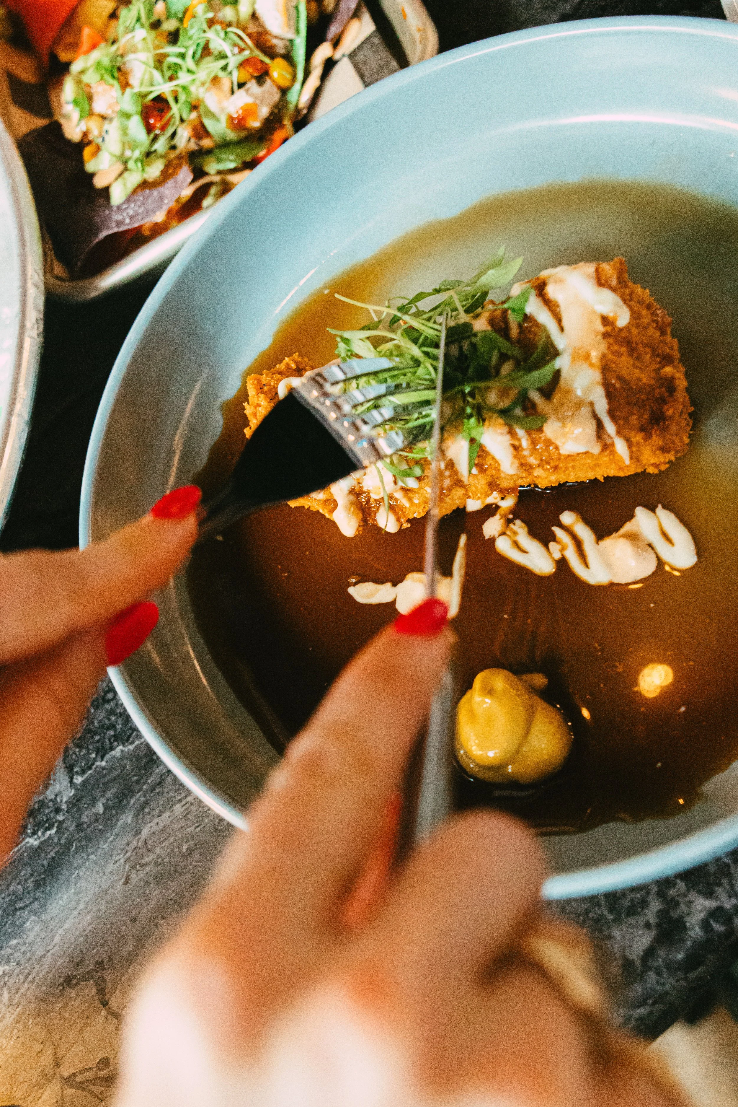 A person holding a fork and knife cutting into a breaded fish fillet topped with microgreens and drizzled with white sauce, served in a bowl with dark soy sauce and a dollop of mustard, alongside a salad in the background.