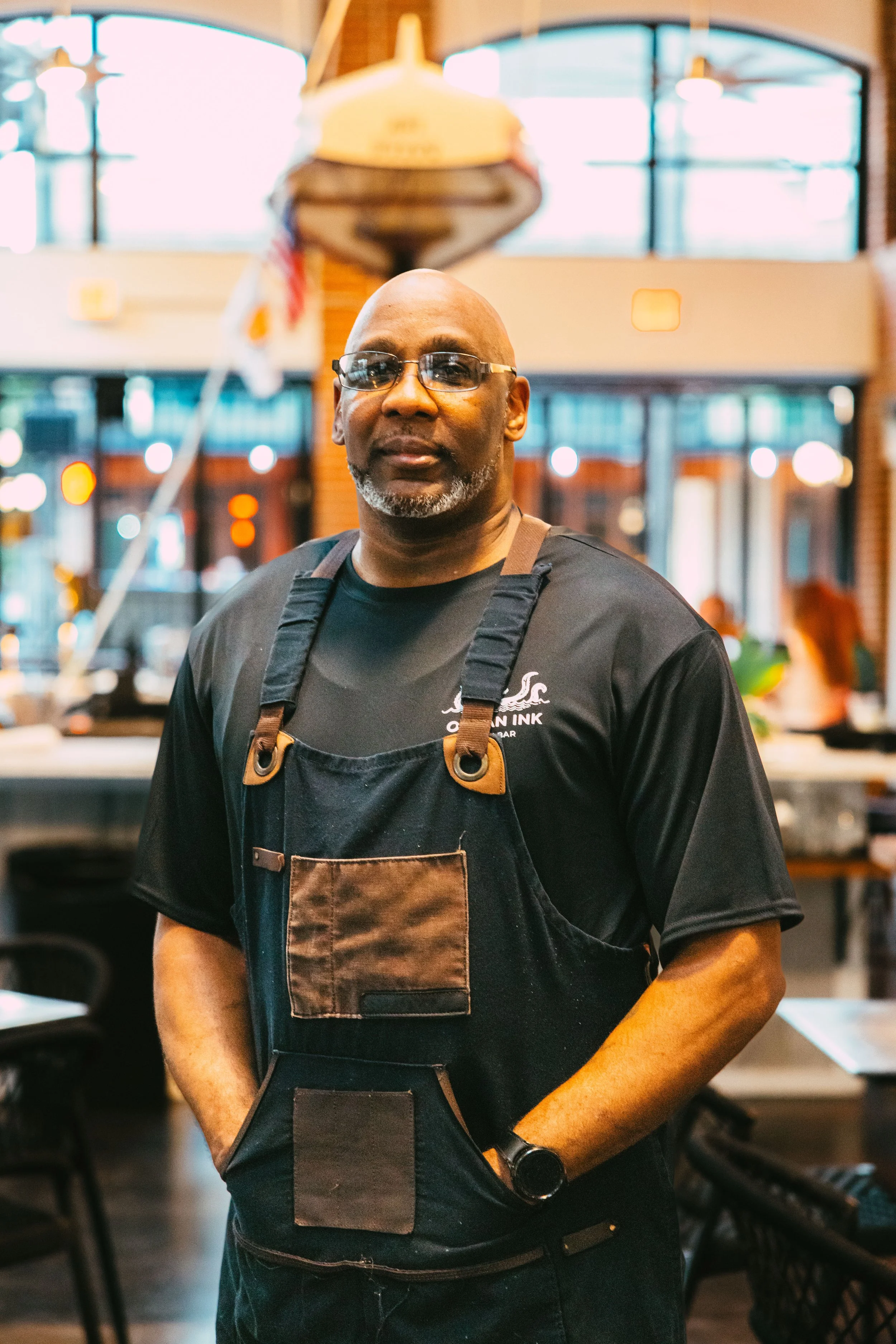 A man wearing glasses, a black t-shirt, and a black apron stands inside a restaurant or café, with blurred background of indoor seating and fixtures.