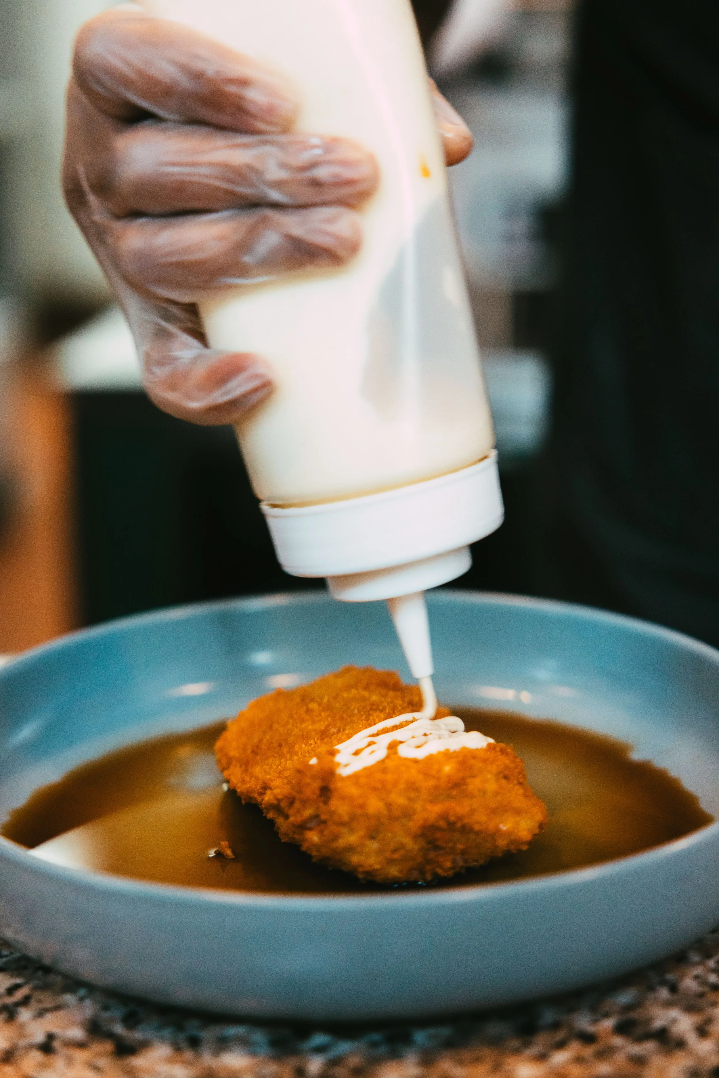 A hand in a plastic glove is squeezing white sauce from a bottle onto a breaded fried cutlet in a blue bowl filled with brown gravy.