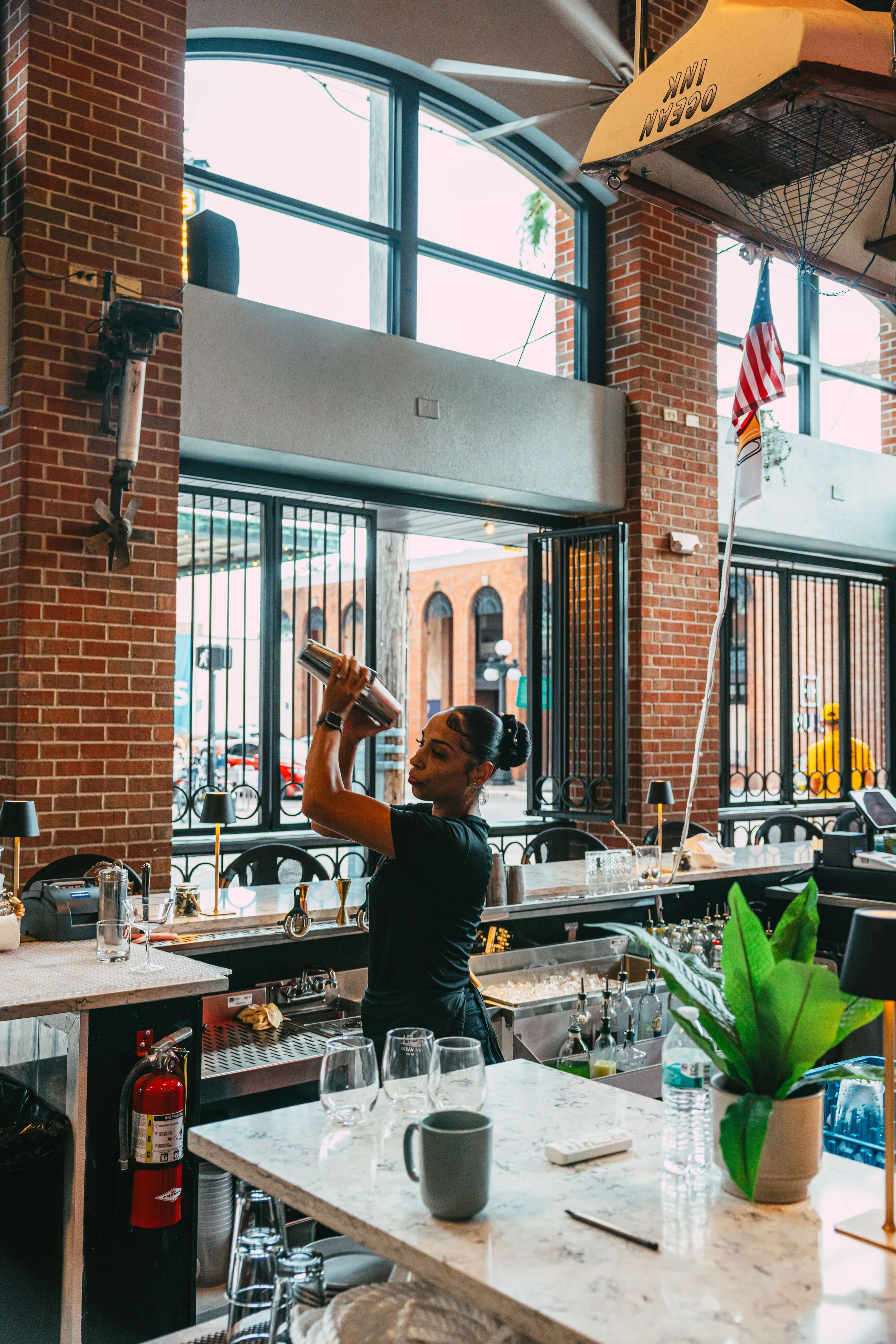 A woman bartender pouring a drink behind a bar in a restaurant with large windows and brick walls.