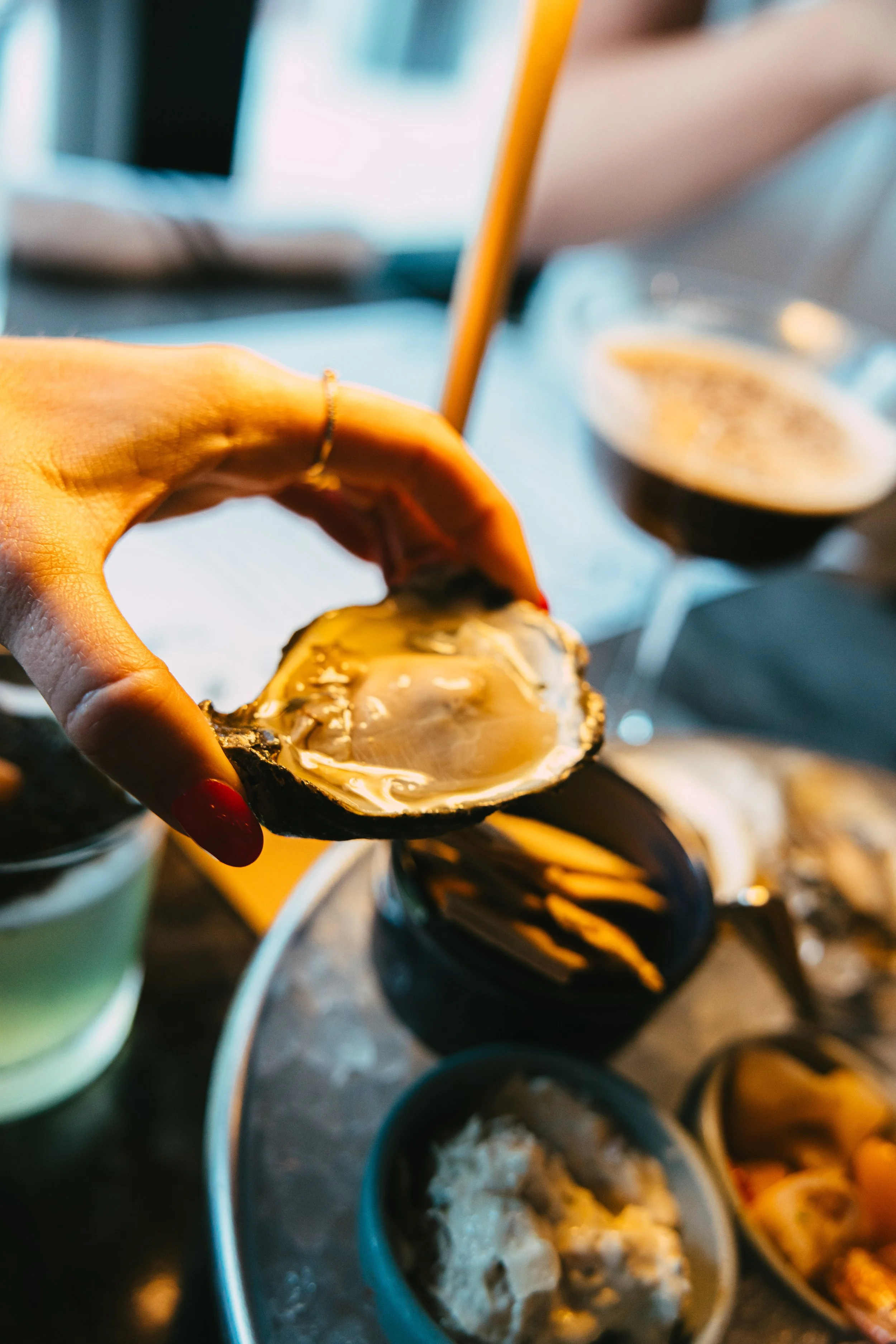 Close-up of a hand holding a freshly opened oyster above a tray with other seafood dishes and drinks.