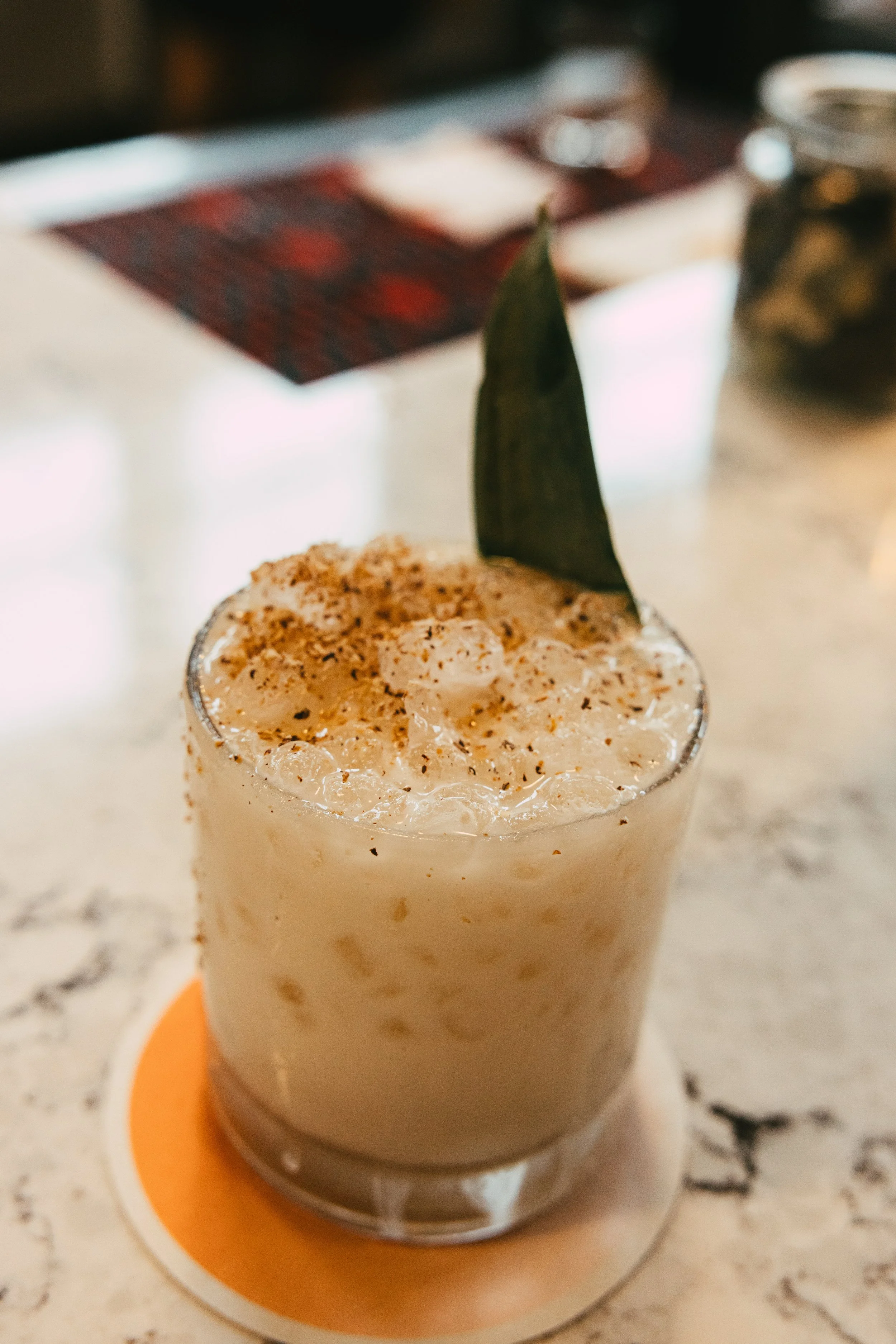 A creamy cocktail topped with crushed ice, a leaf garnish, and sprinkled with spices on a clear glass, placed on a coaster on a marble countertop.