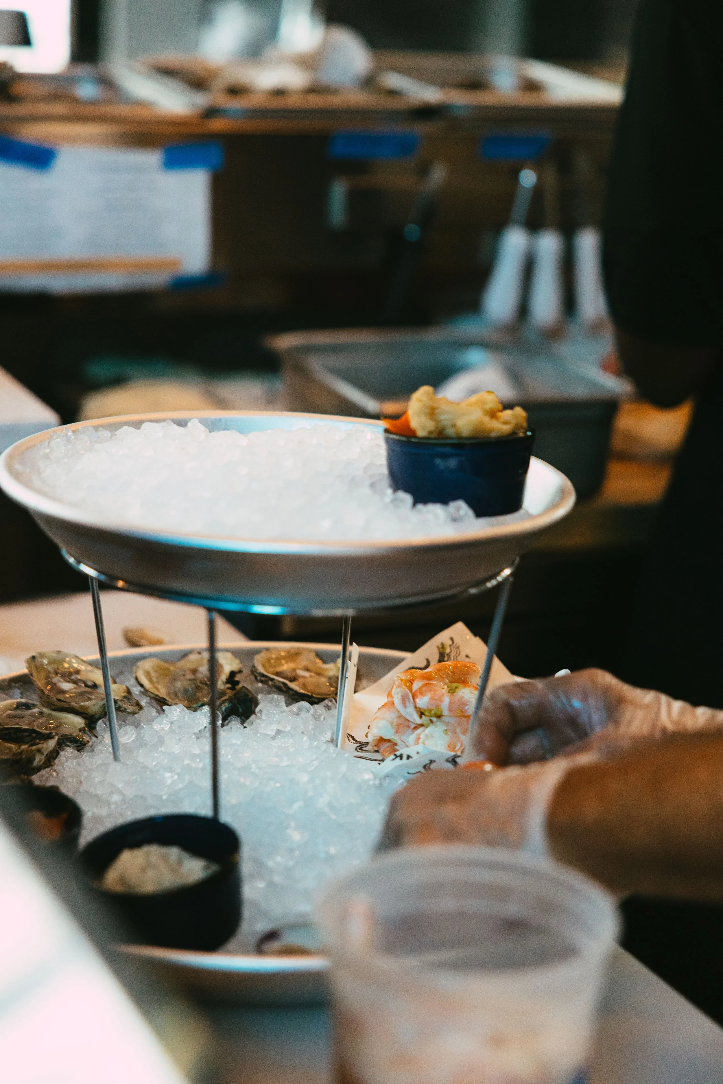A seafood display with oysters on ice on a tiered tray, a small bowl of sauce, and a hand reaching for seafood in a restaurant or market setting.