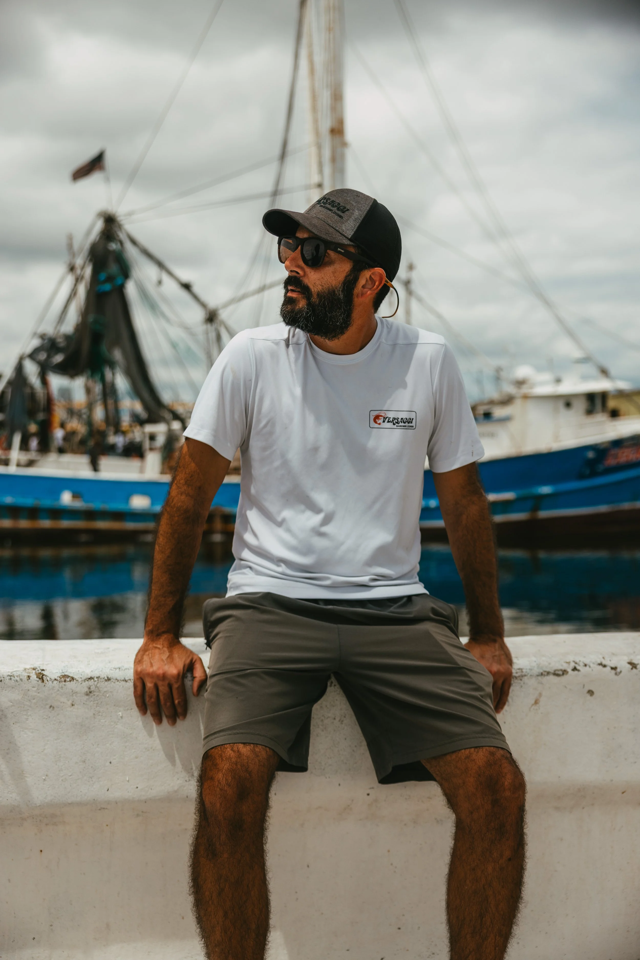 Man with beard wearing sunglasses, a cap, a white T-shirt, and shorts sitting on a white wall near a marina with sailboats and cloudy sky in the background.