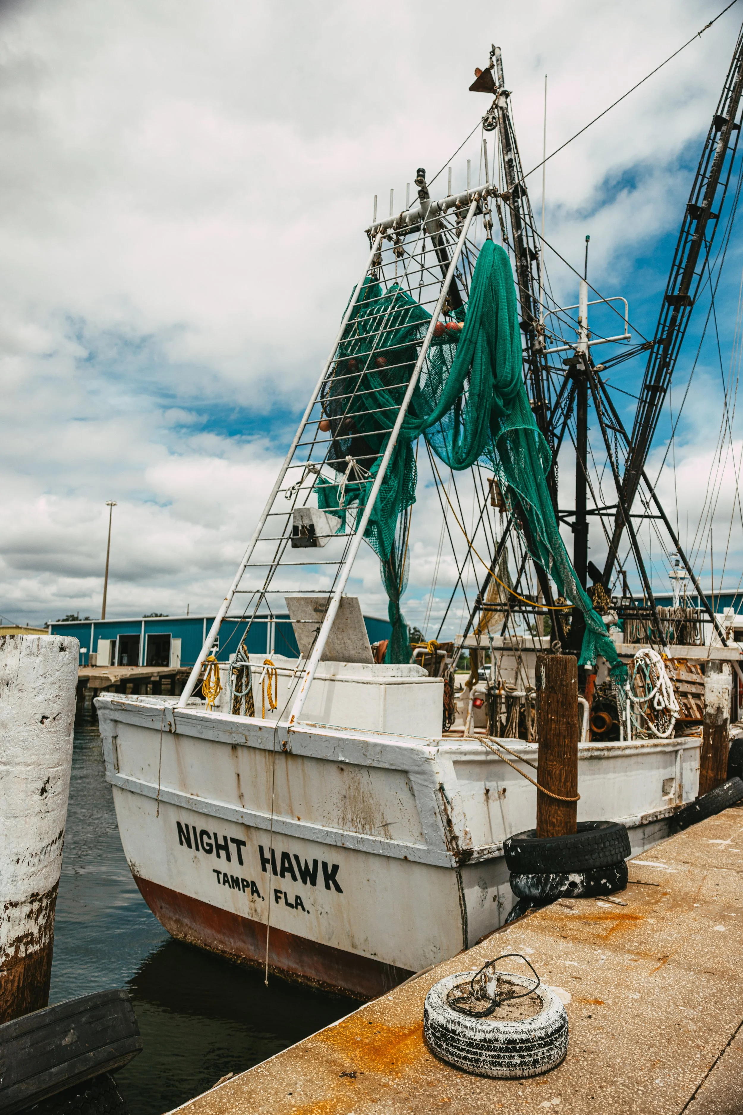 A fishing boat named 'Night Hawk' docked at a pier in Tampa, Florida, with fishing nets hanging on the boat and a cloudy sky overhead.