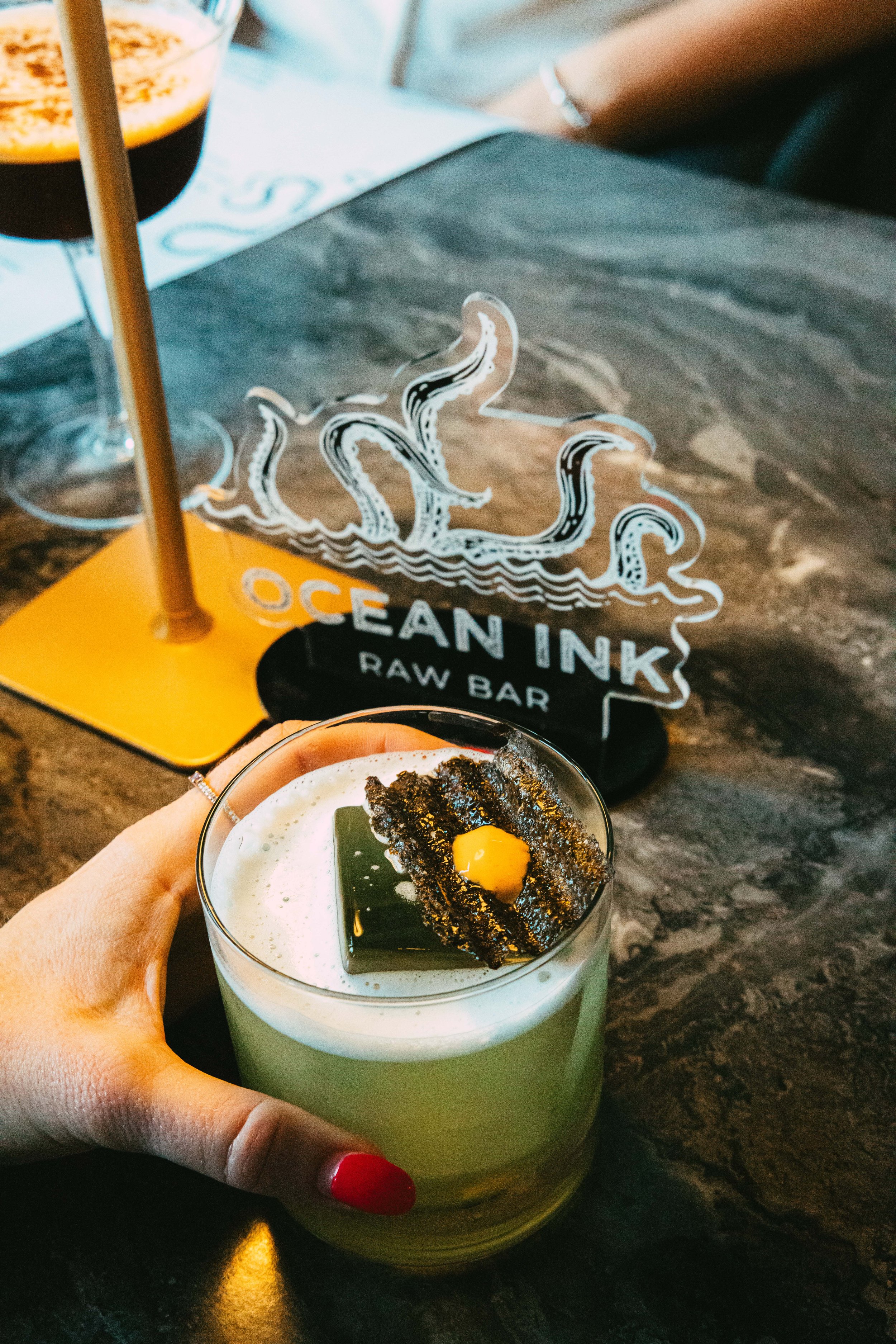 A hand holding a glass of green smoothie topped with a piece of seaweed, a yellow egg yolk, and dark-colored crumbles, sitting on a dark marble table. In the background, there is a sign that says 'OCEAN INK RAW BAR' with an octopus illustration and t