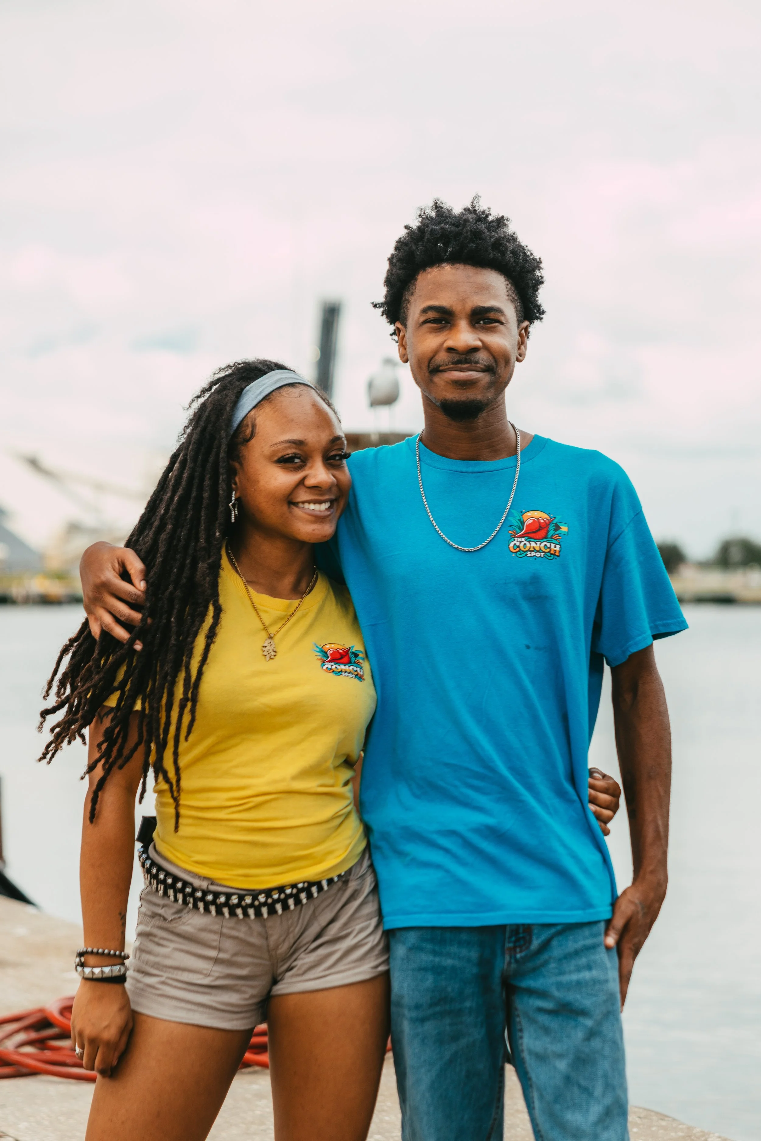 Two young adults, a woman with dreadlocks and a man with curly hair, standing close by a body of water, smiling at the camera. They are wearing colorful t-shirts with a logo, indicating they are part of a group or event, with the woman in yellow and 